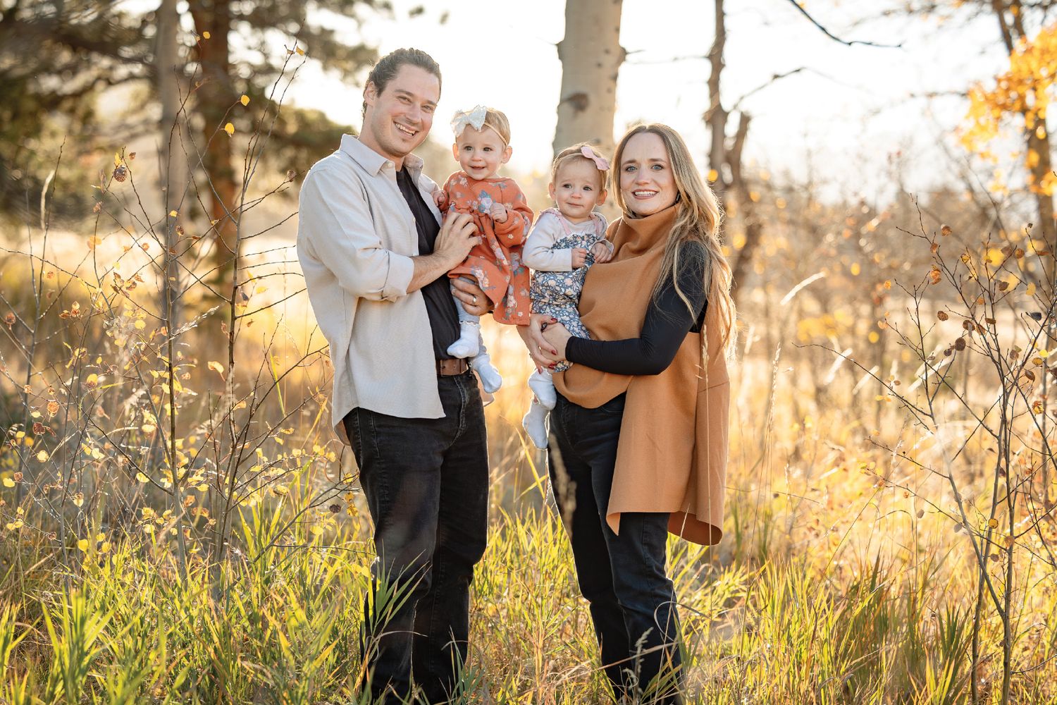 Family of four poses outdoors in golden autumn light, parents holding two toddlers among tall grass and trees.