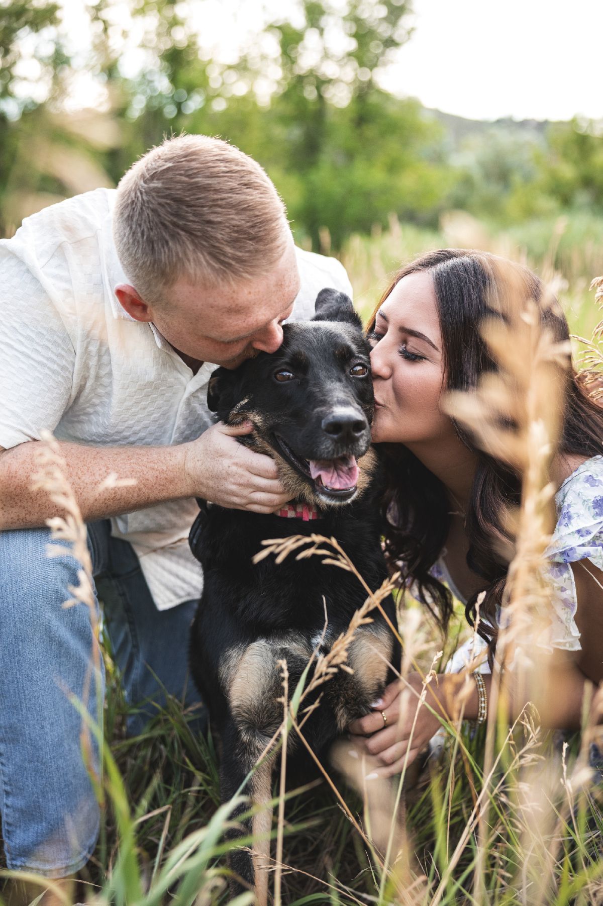 Couple kissing their happy black dog in a lush green field, capturing a sweet outdoor pet photography moment.