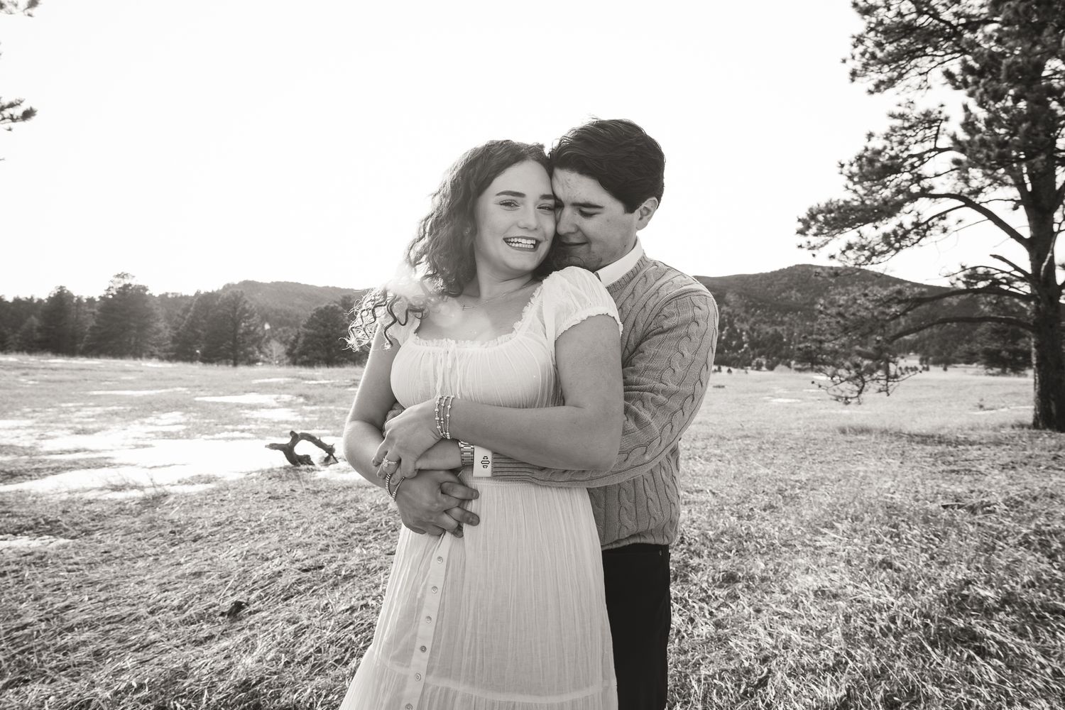 Black and white photo of a couple embracing in a meadow, woman in white dress, man in sweater, with trees and hills behind.
