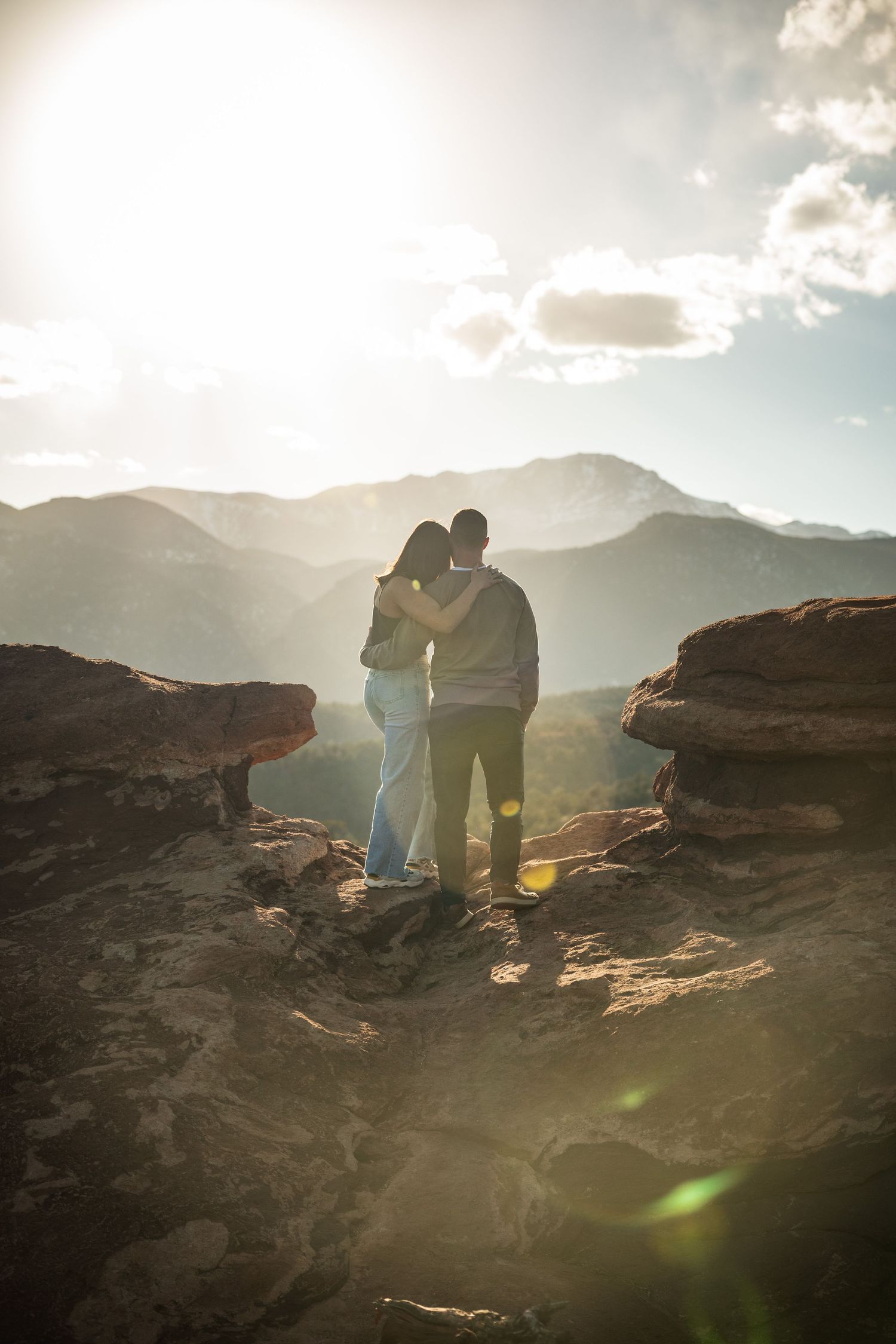 Couple overlooking mountain vista at sunset while standing on rocky outcrop.