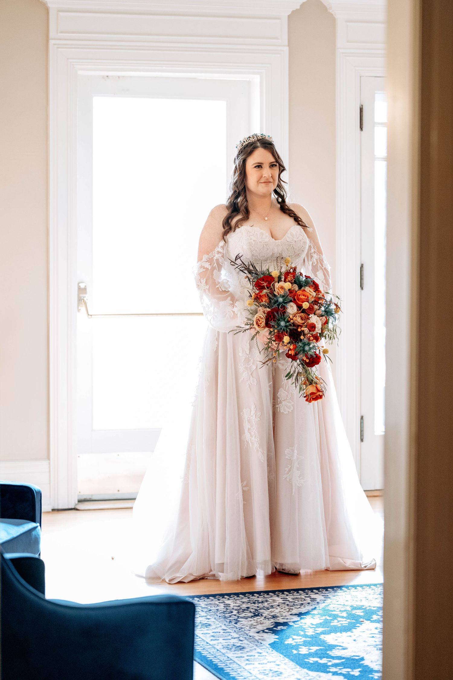 A bride in a flowing white lace gown holds a cascading bouquet of coral and orange flowers in an elegant doorway.