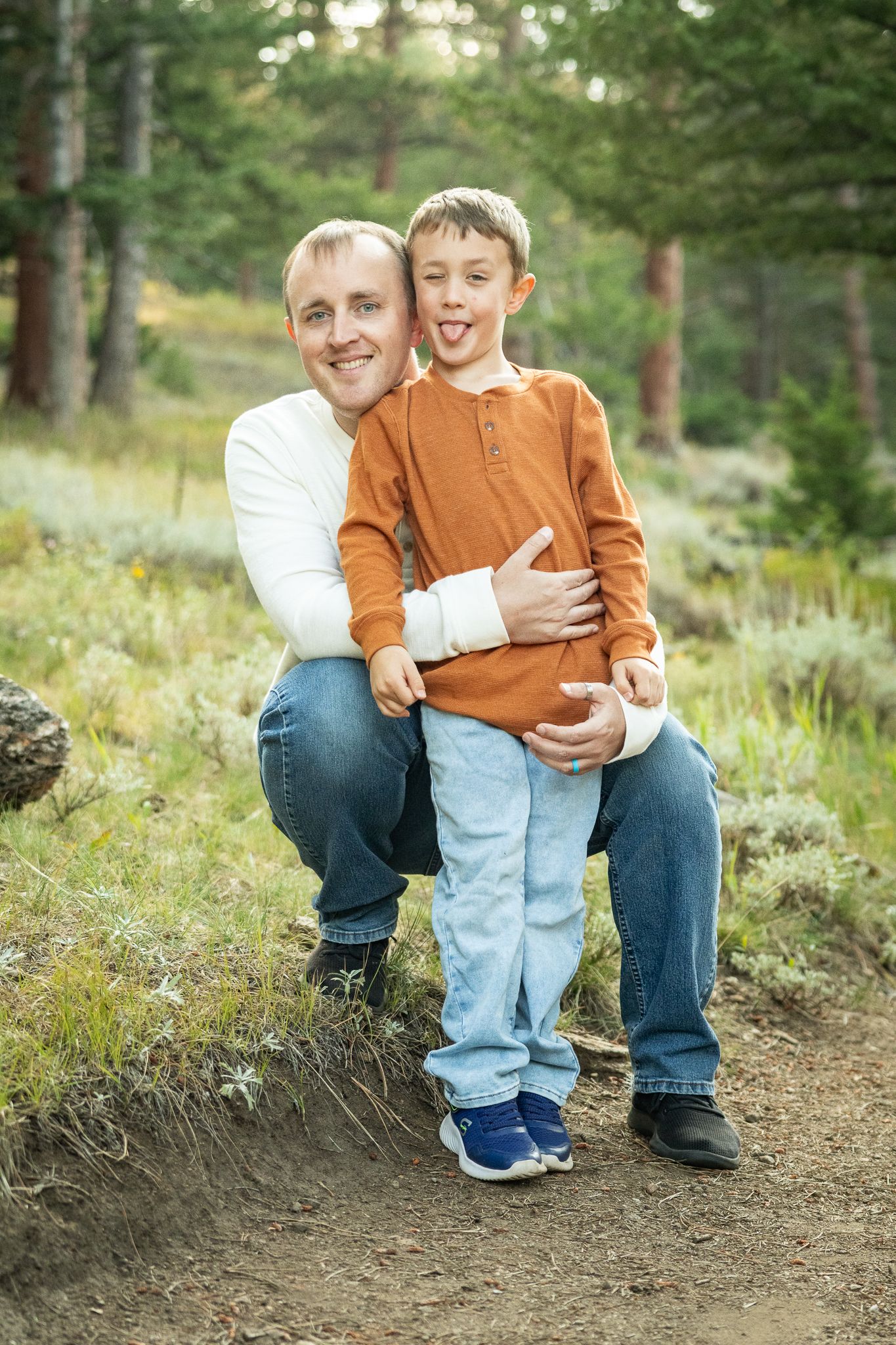 Father and young son smiling together on a forest trail, boy sticking out tongue playfully in outdoor family portrait.