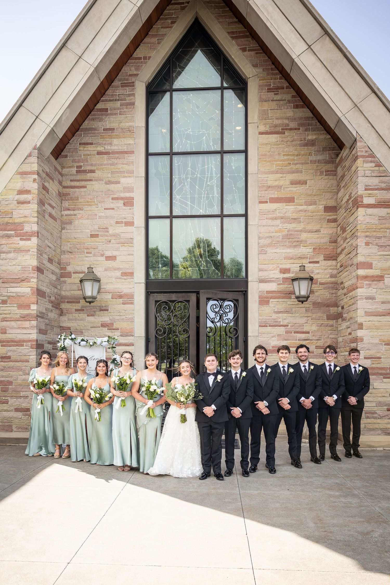 Wedding party poses outside brick chapel, bridesmaids in sage green gowns and groomsmen in black tuxedos.