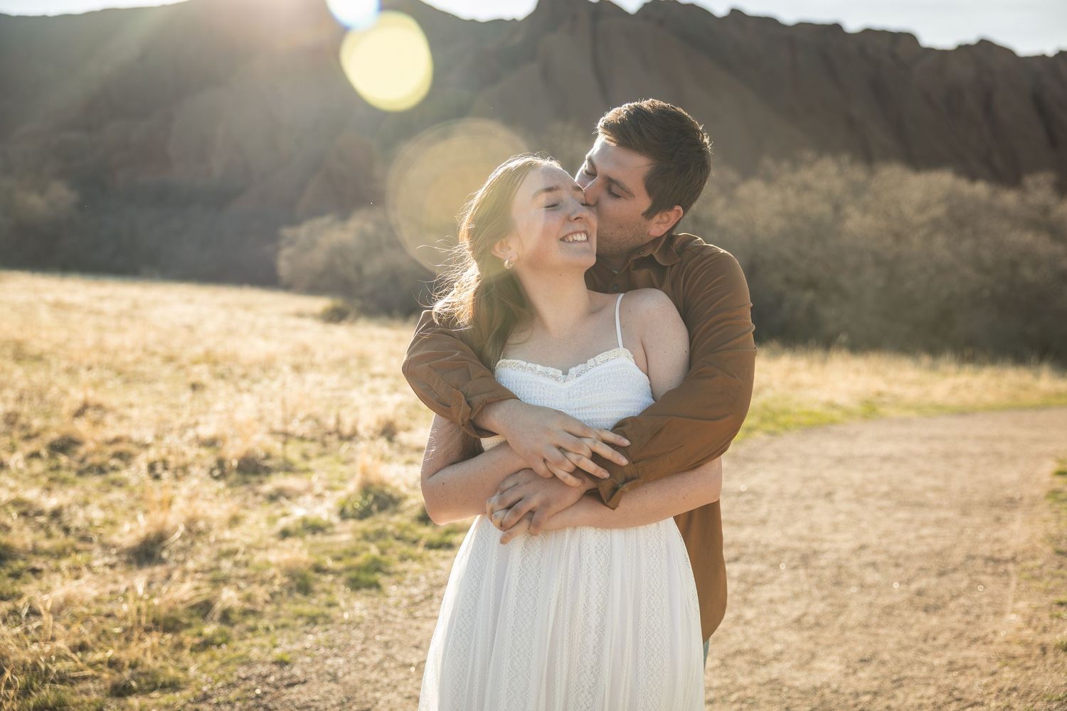 Couple embracing and smiling at golden hour with sunlit rocky mountains in the background.