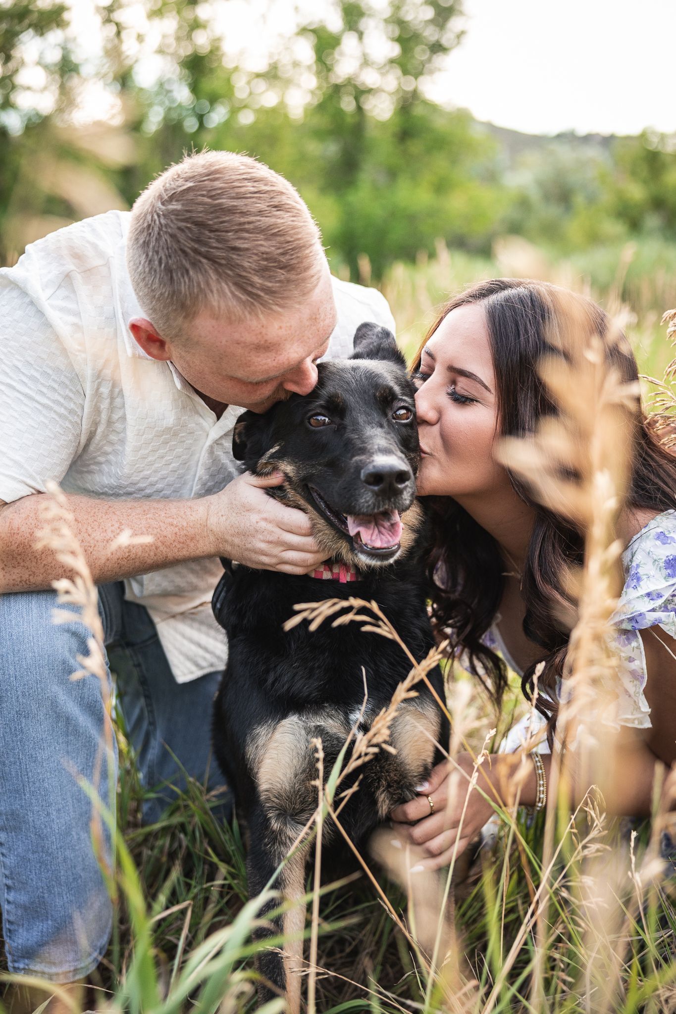A couple kisses their happy black dog in a lush green field during a romantic outdoor photo session.