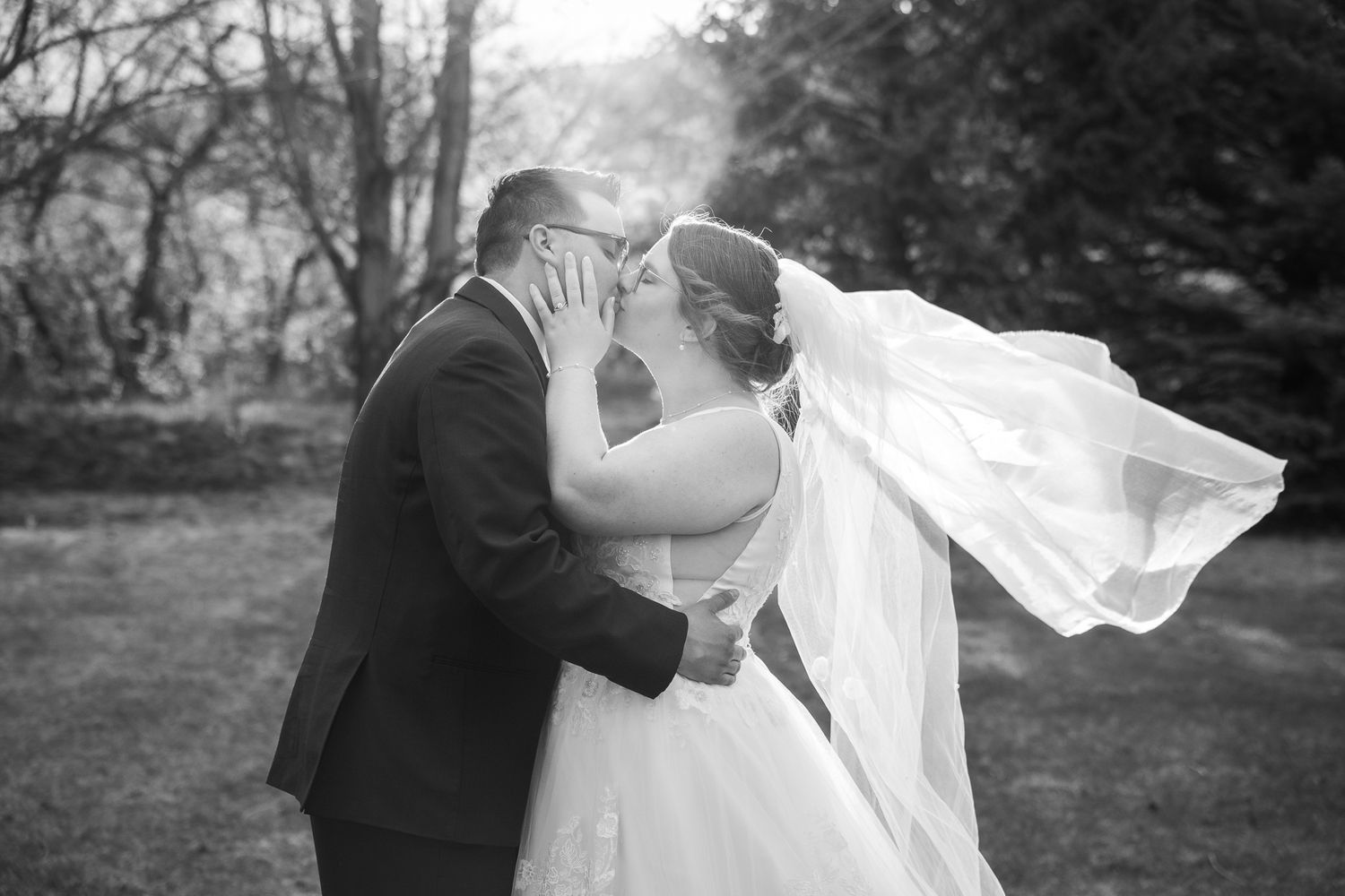 Black and white wedding photo of couple kissing outdoors, bride's veil flowing in wind, surrounded by trees.