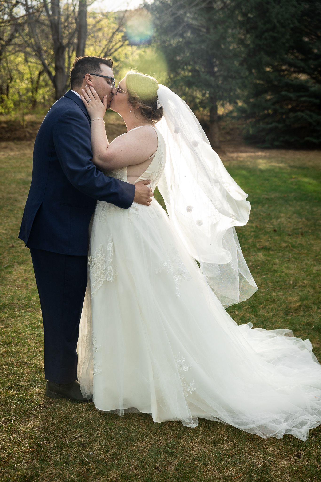 Bride and groom share a romantic kiss outdoors, bride wearing white gown with veil, groom in navy suit.