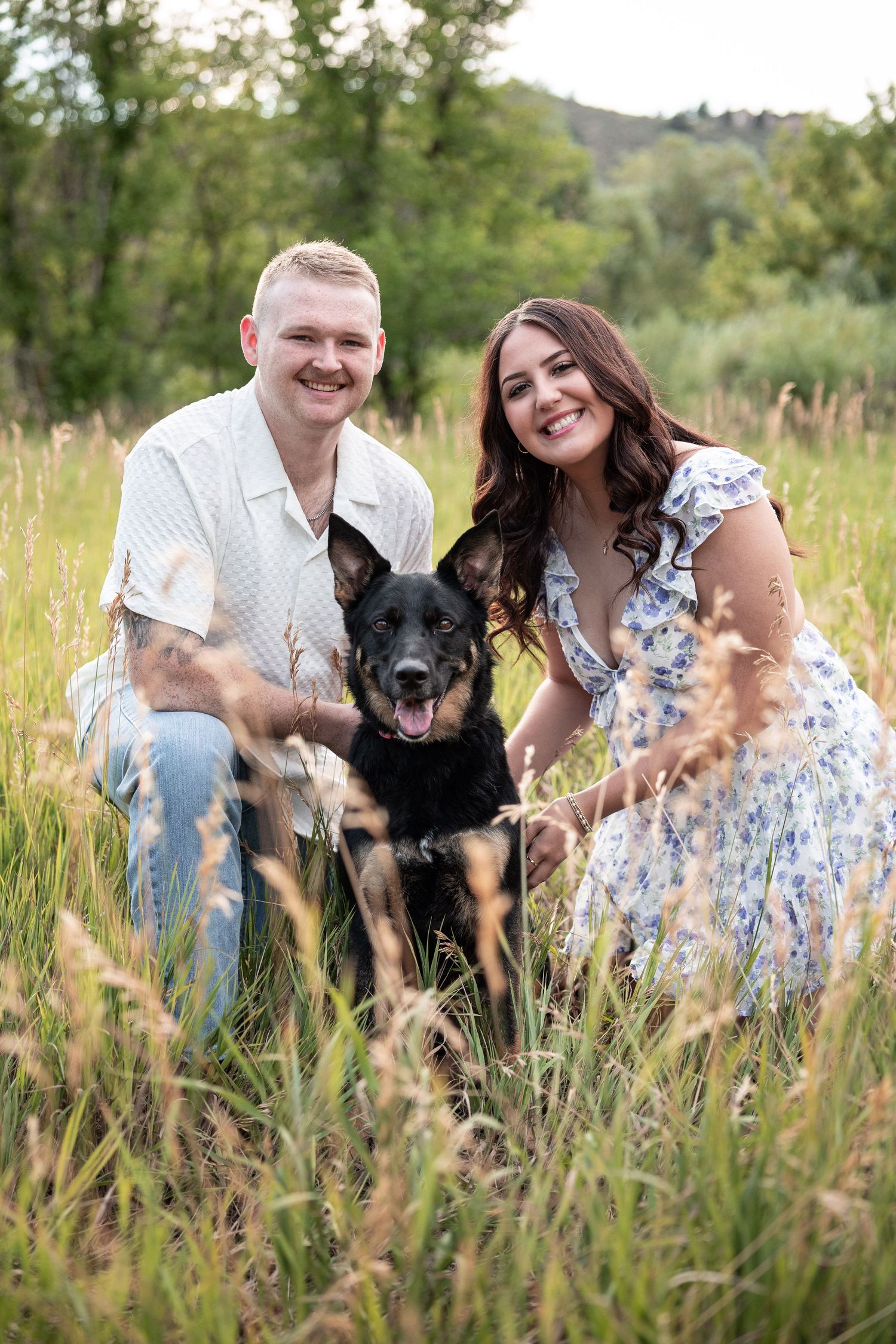 A smiling couple poses with their black German Shepherd dog in a lush green meadow field.