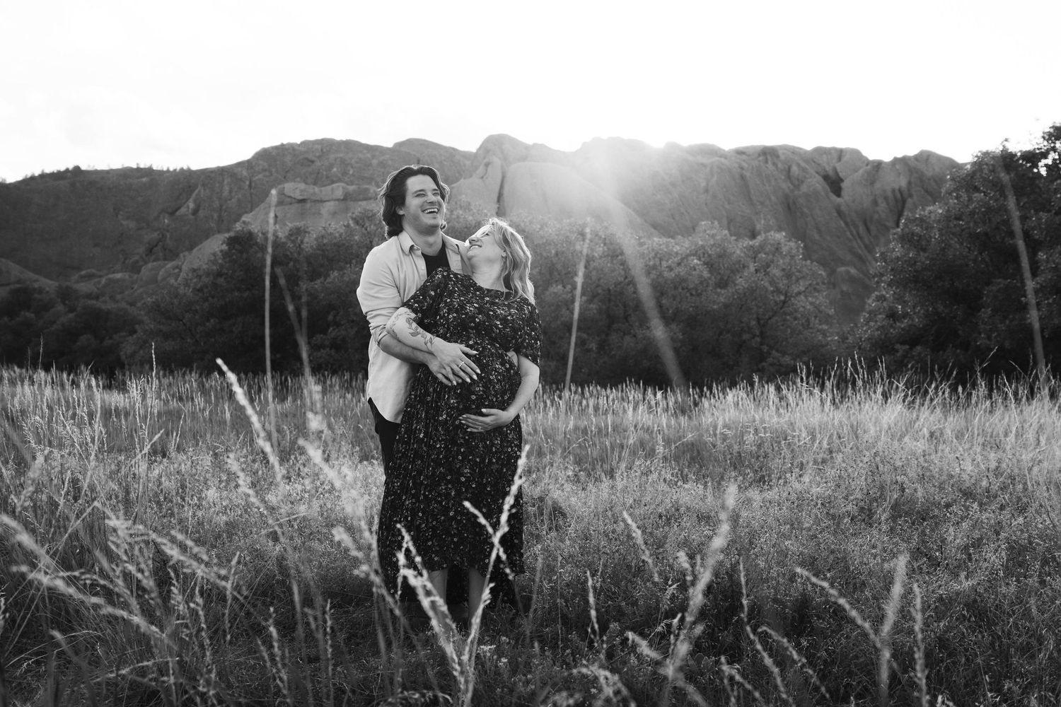 A black and white portrait of a couple embracing in a field at sunset with mountains behind them.