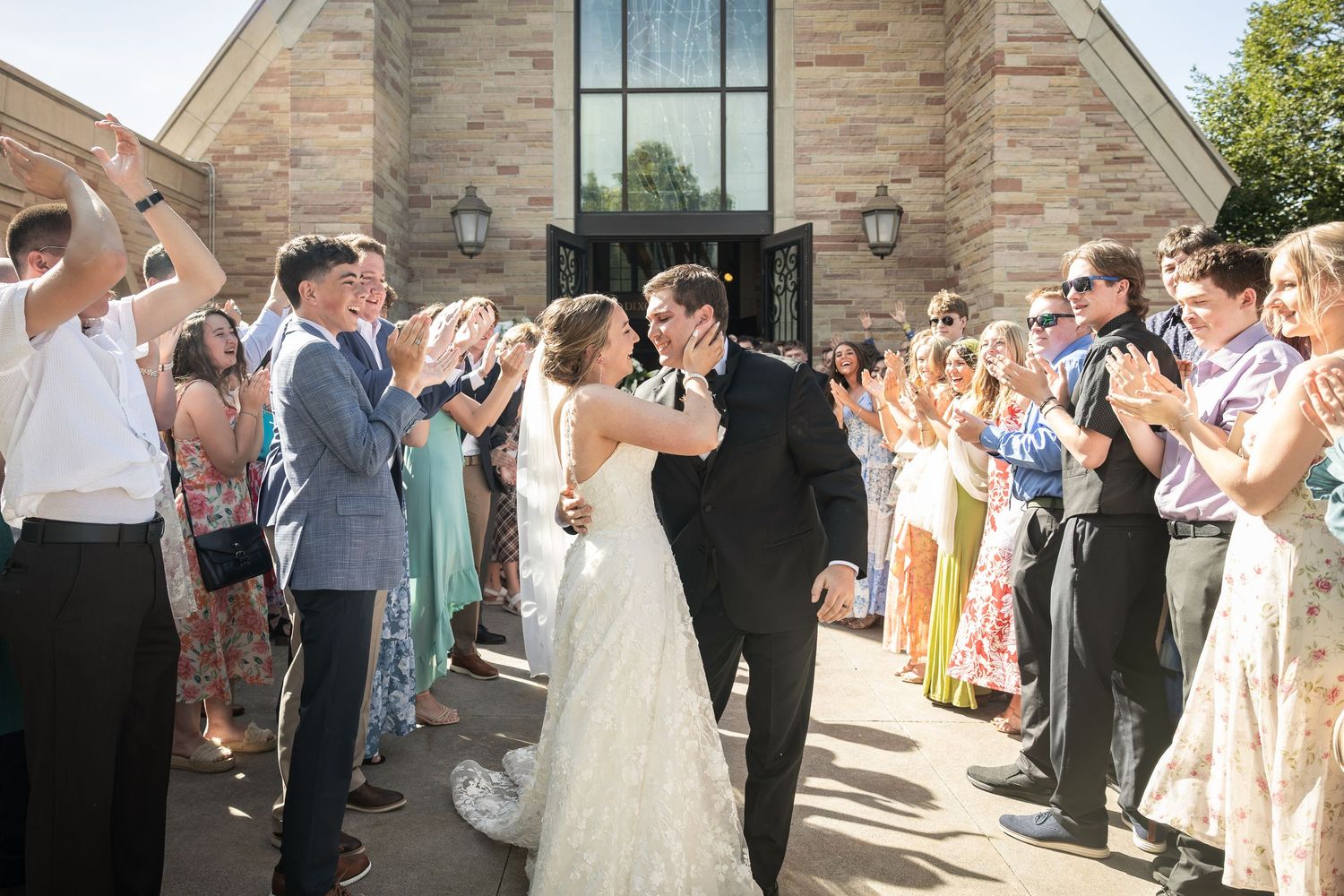 Bride and groom share a kiss outside a stone church surrounded by cheering wedding guests on a sunny day.