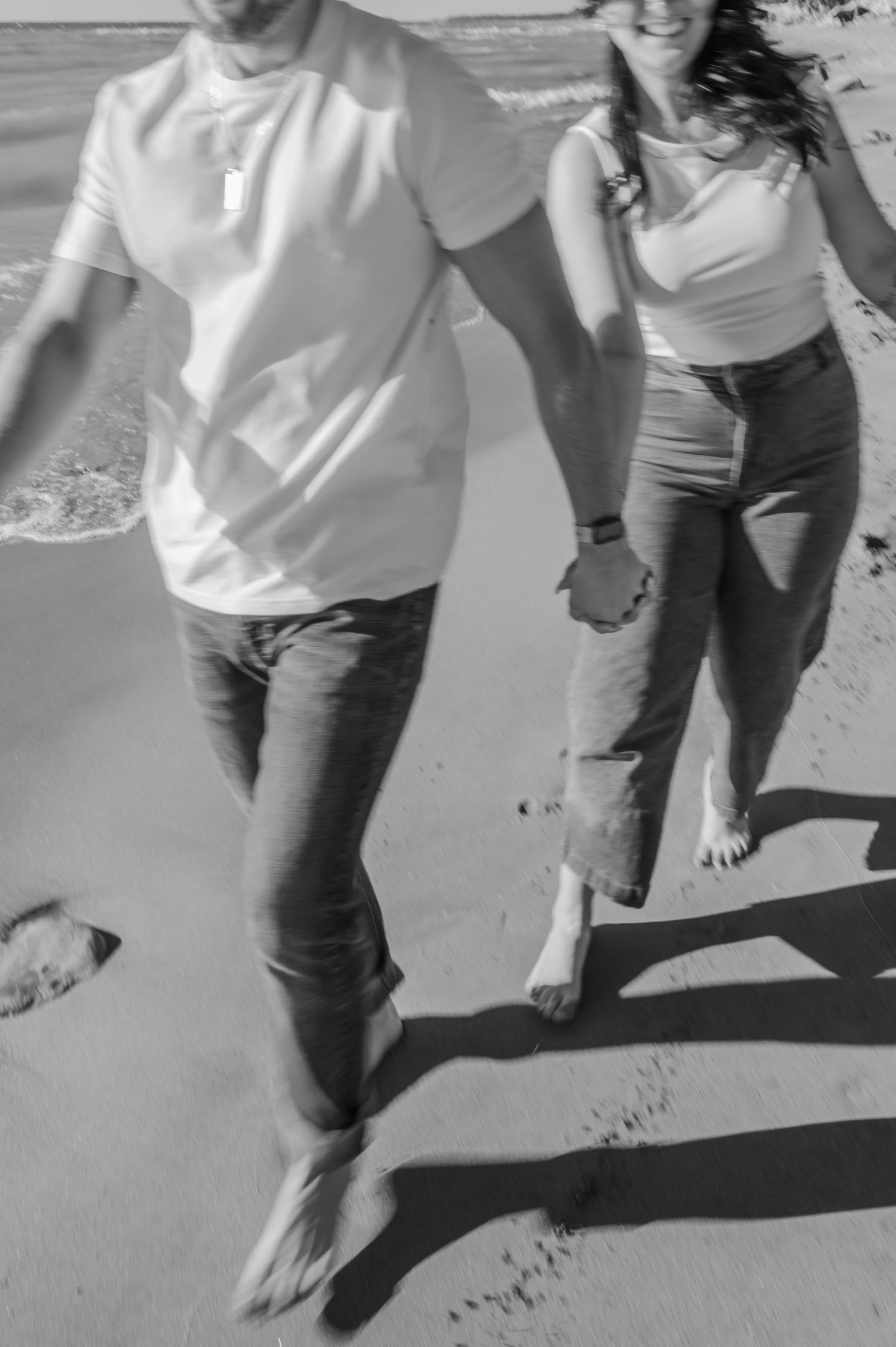 A couple walking barefoot on the beach together holding hands in black and white photography.