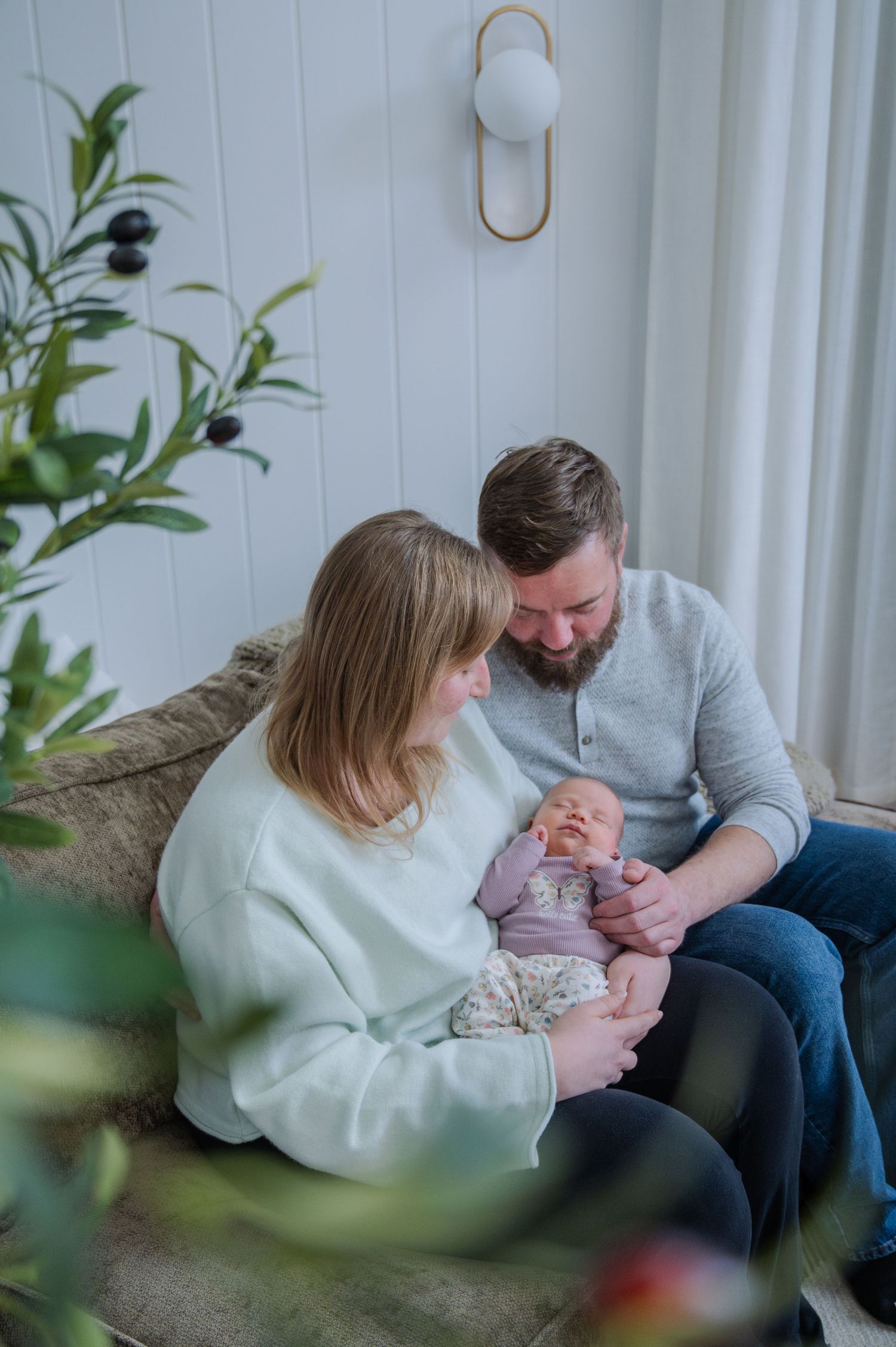 A couple sits on a couch admiring their newborn baby in a bright, cozy living room with green plants nearby.