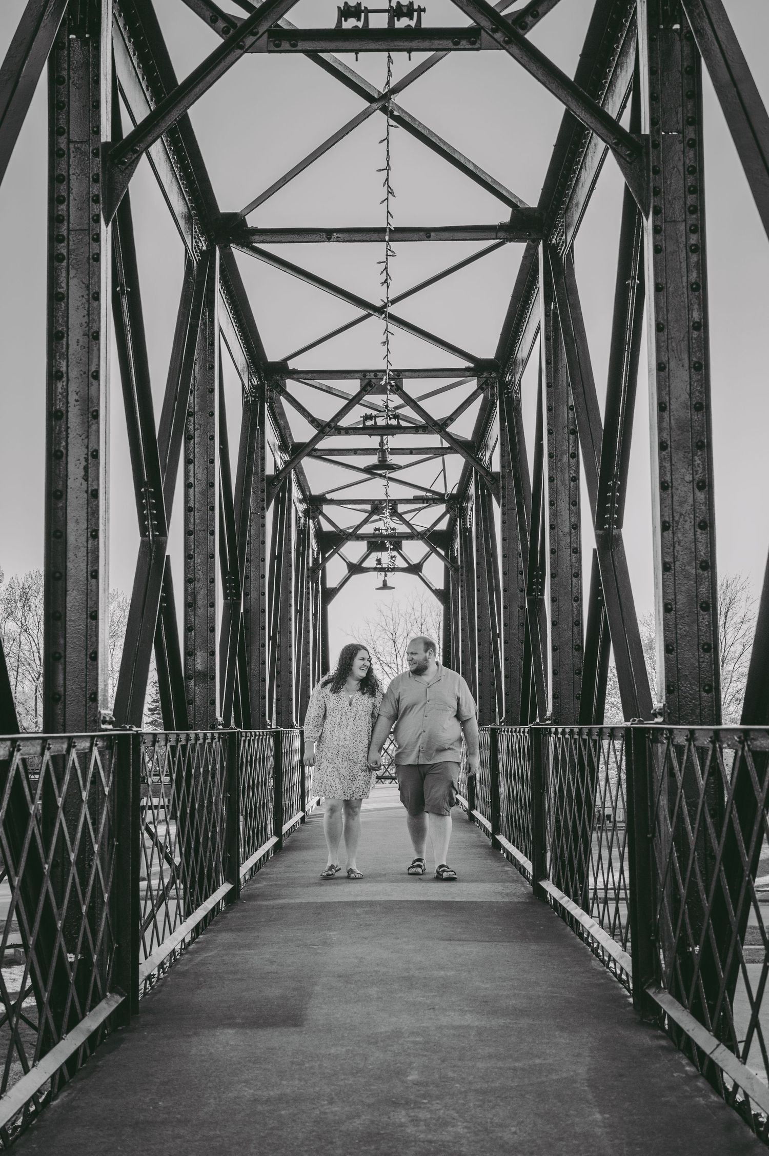 A black and white photo of a couple walking along a metal bridge with geometric industrial architecture.