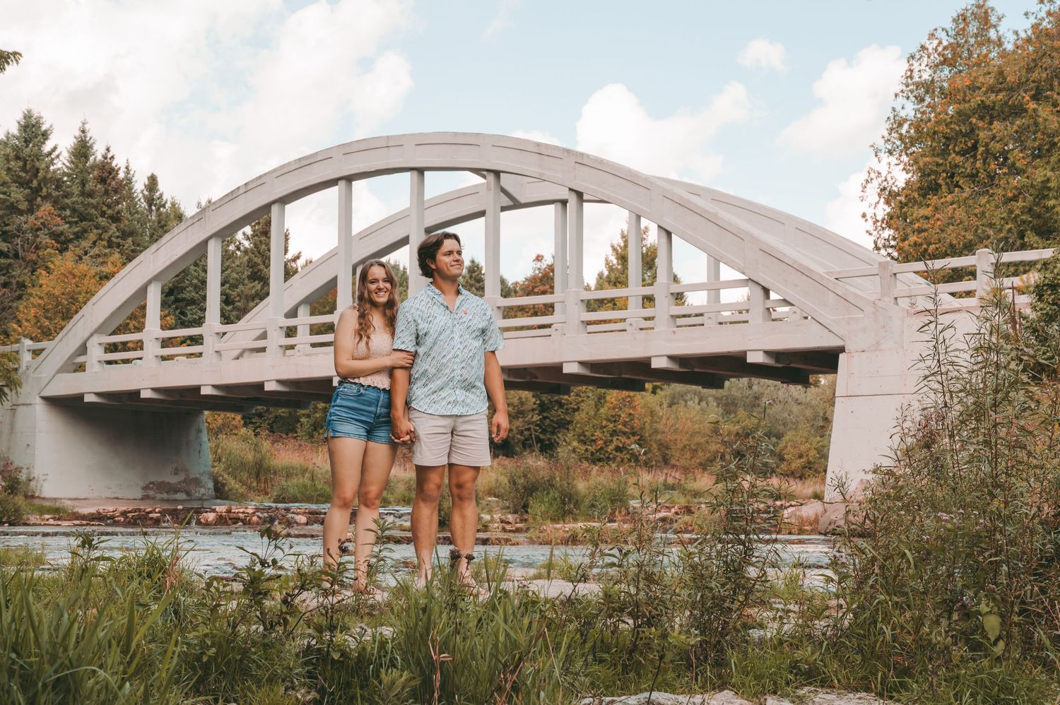 Couple posing together on rocky riverbed with white arched bridge in background.