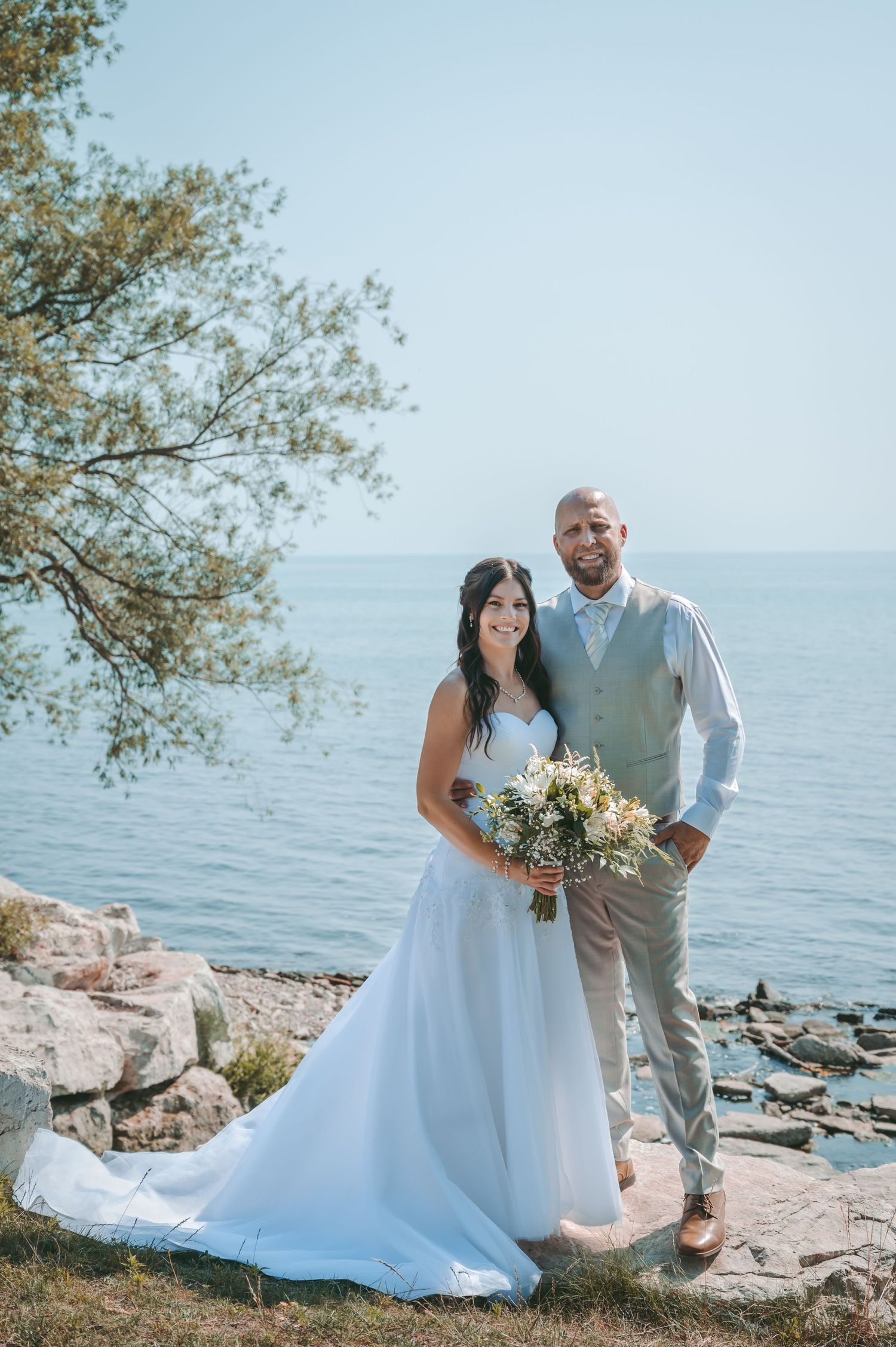 Coastal wedding portrait featuring couple standing on rocky shoreline with water view.