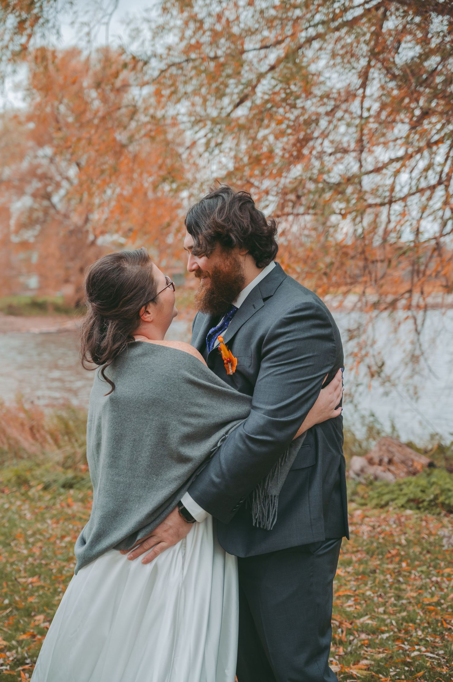 A romantic autumn wedding portrait showing a tender embrace between newlyweds by a lake with fall foliage in the background.