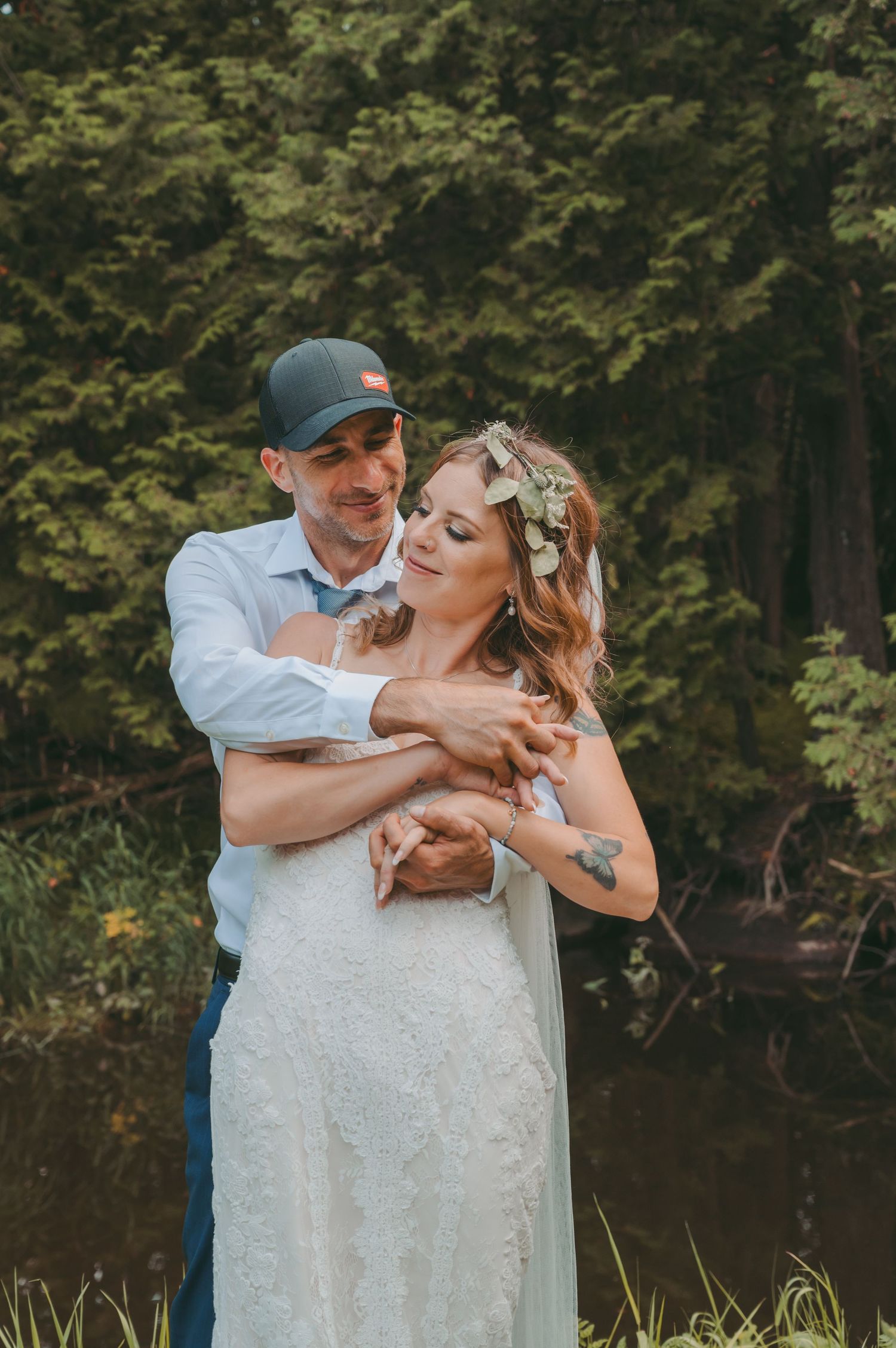 A wedding couple embraces outdoors, with the bride wearing a white dress and floral crown.