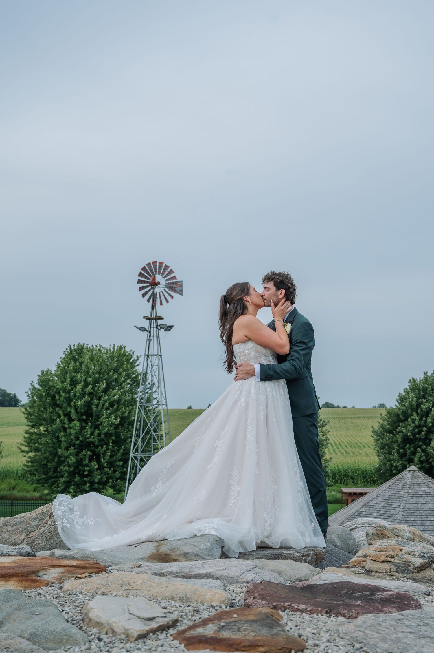 Bride and groom share a kiss on stone patio with rustic windmill and green fields backdrop on their wedding day.