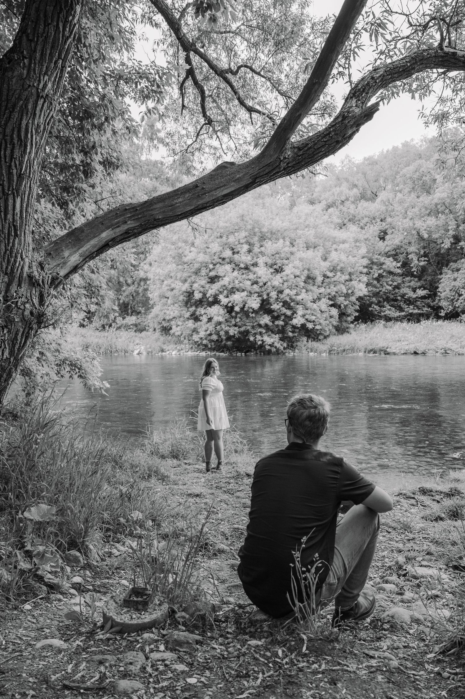 Black and white photo of a man sitting by a river watching a woman in a white dress standing near the water's edge.