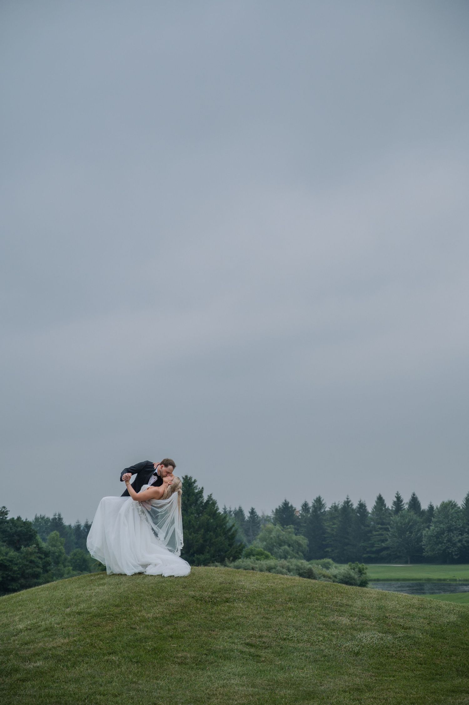 Groom dips bride in white gown on a lush green hill with evergreen trees and overcast sky backdrop.