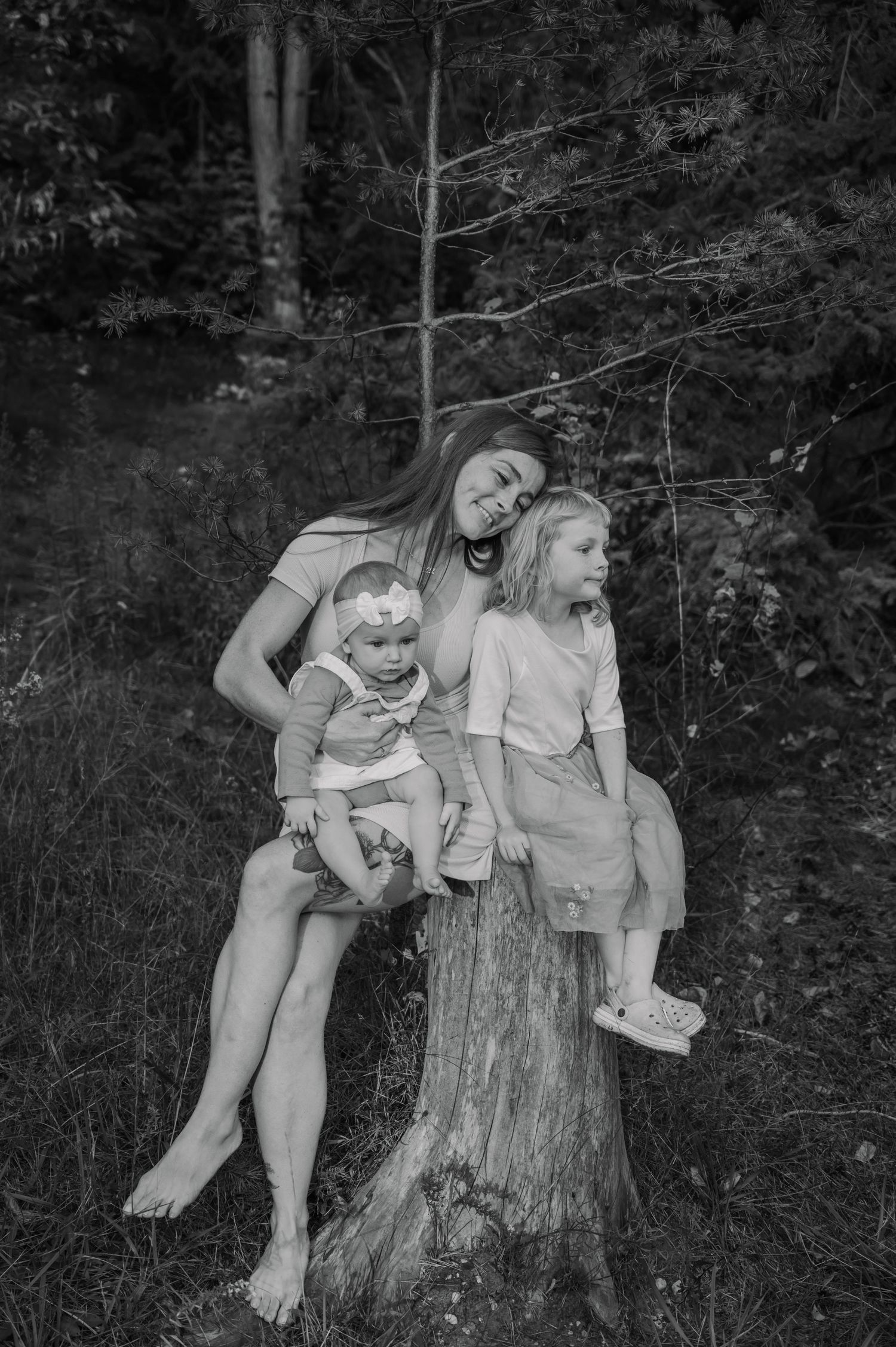 Mom and daughters sit together on a tree stump in a wooded area, sharing laughter and conversation.