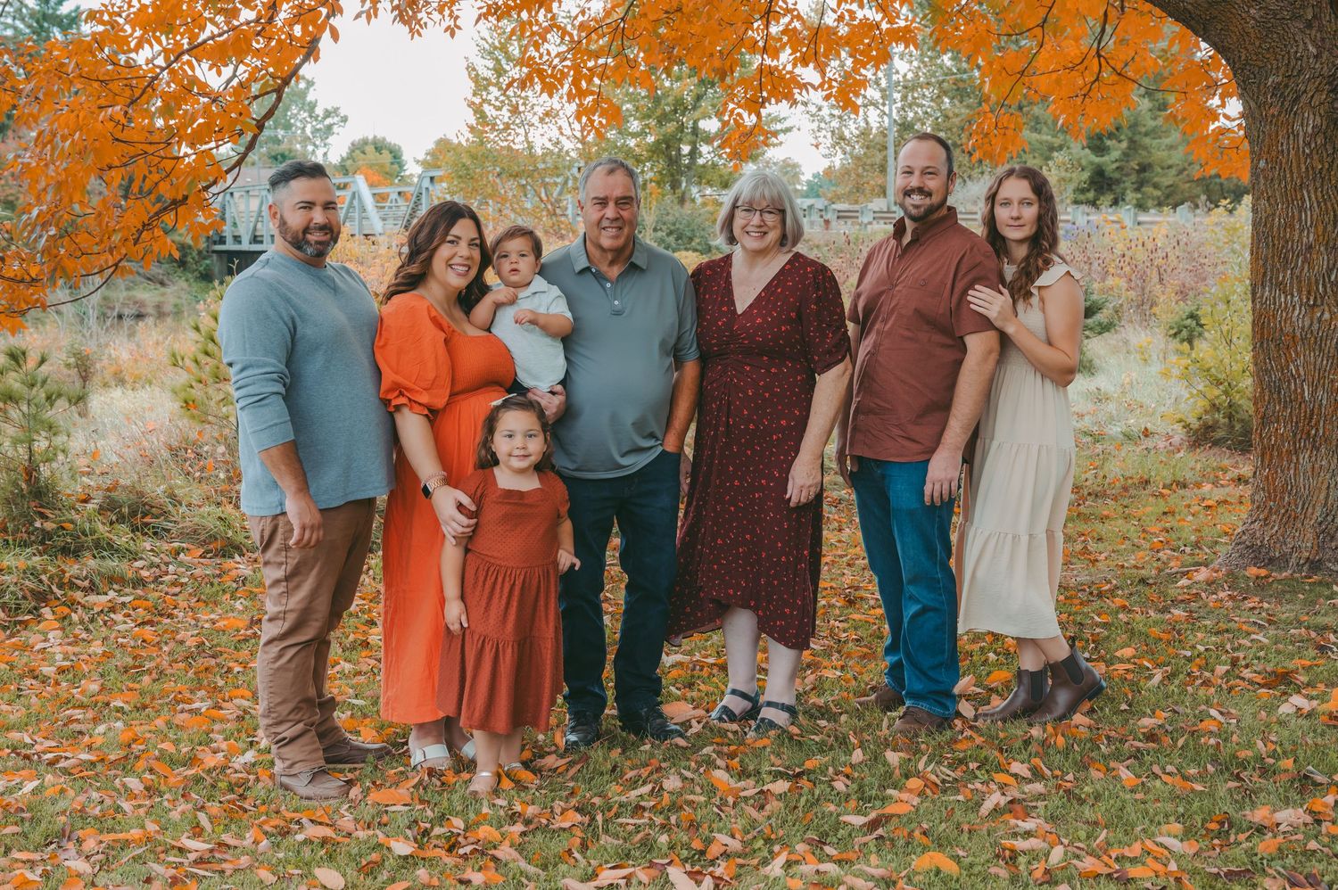 Multi-generational family portrait under vibrant orange autumn trees wearing coordinated fall colors.