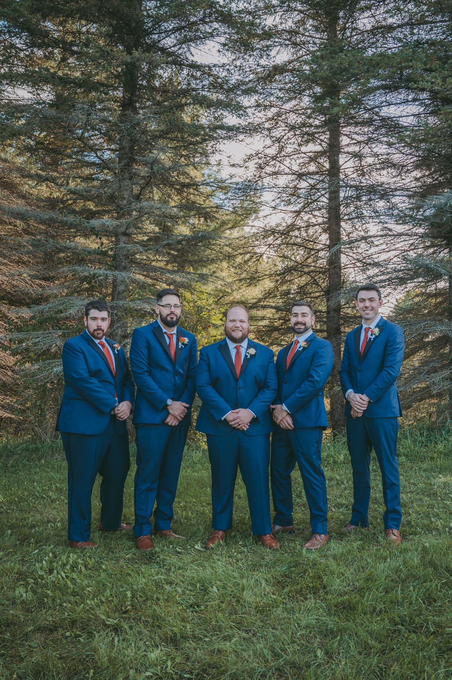 Groomsmen in matching navy suits with orange accents pose in front of pine trees.