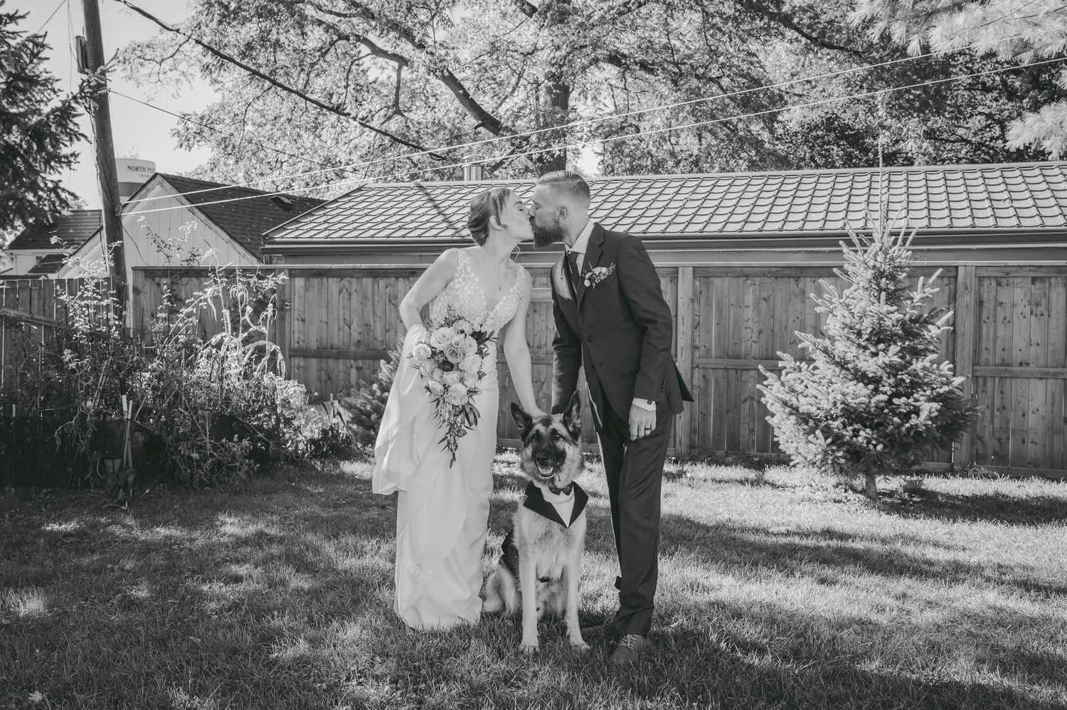 A vintage looking wedding photo outdoors with a dog joining the couple in front of wooden buildings.