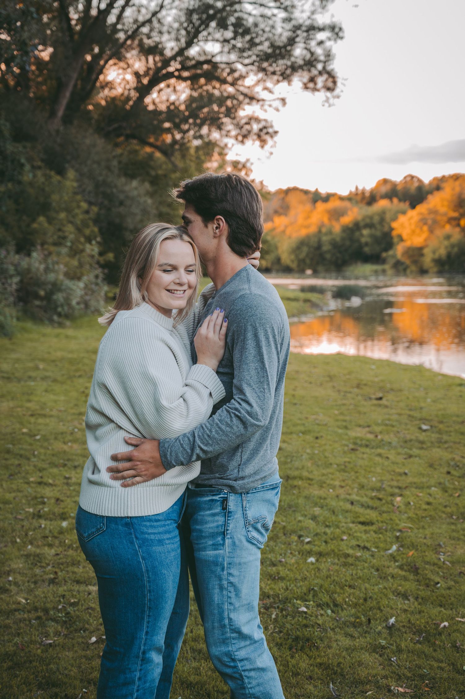A couple shares an intimate embrace on grass with fall colors and a lake in the background.