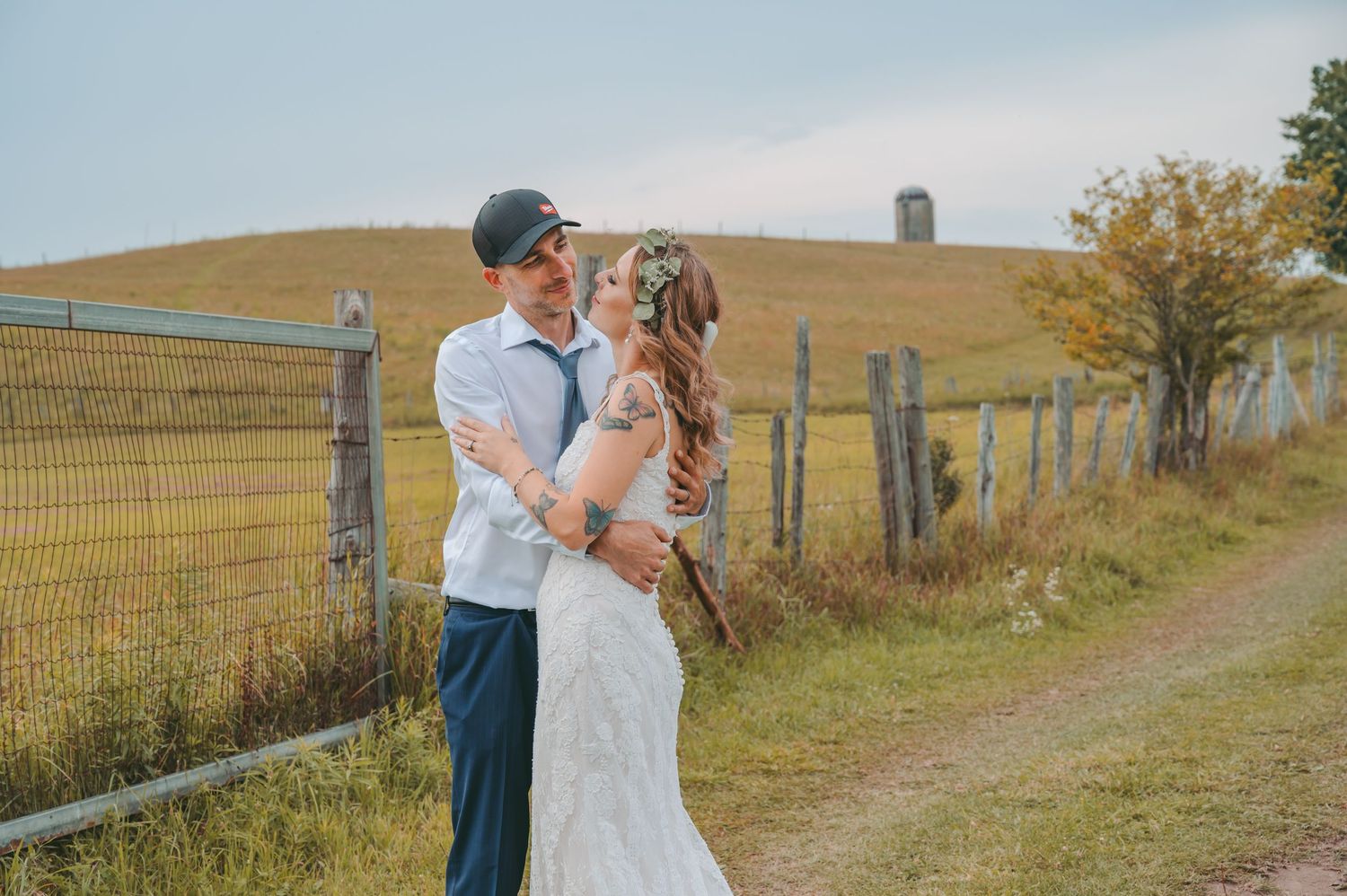 Romantic couple embraces in a scenic countryside setting with golden rolling hills and wooden fences in the background.