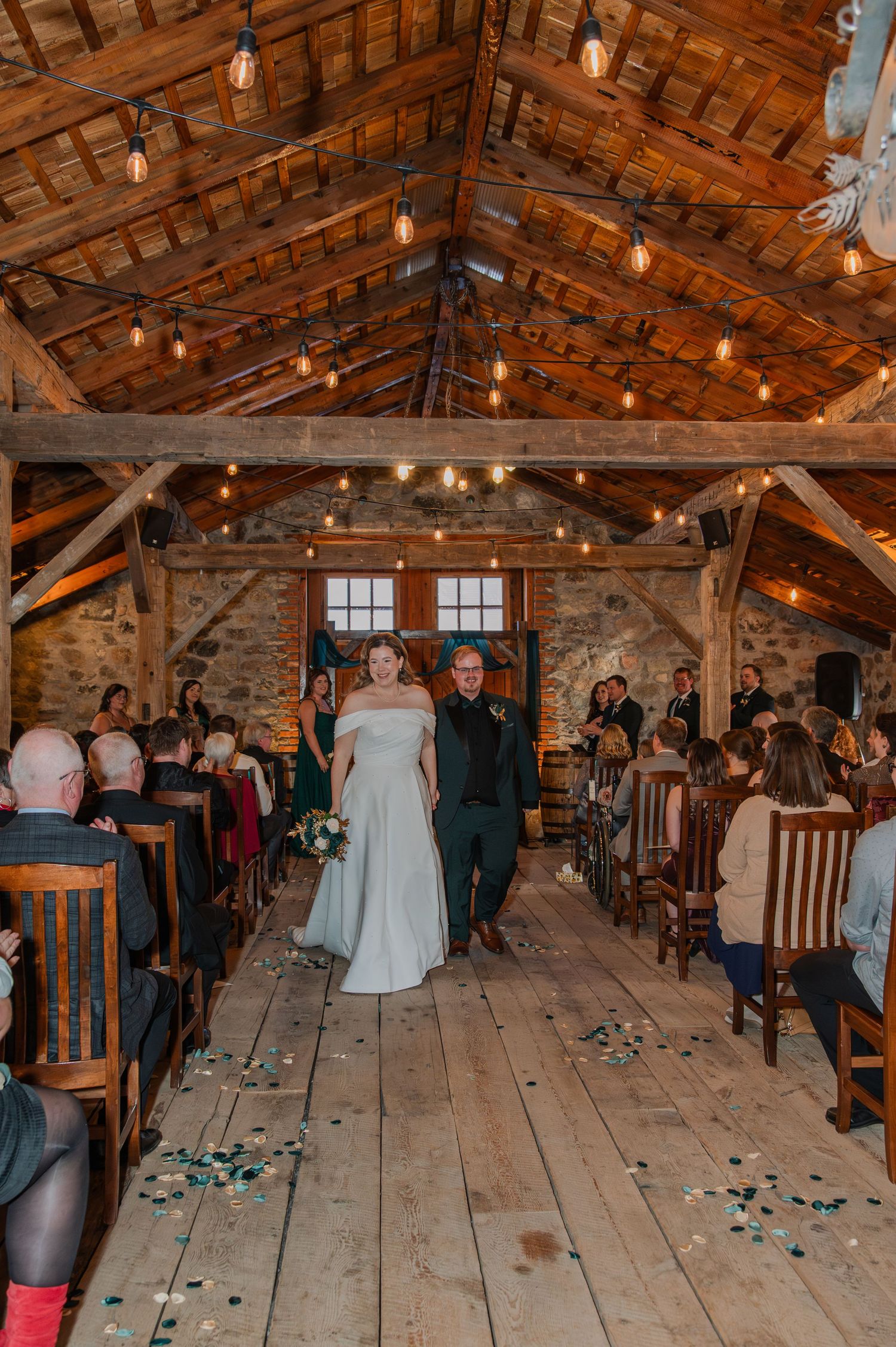 Rustic barn wedding ceremony with wooden beams, string lights, and scattered petals on the floor.