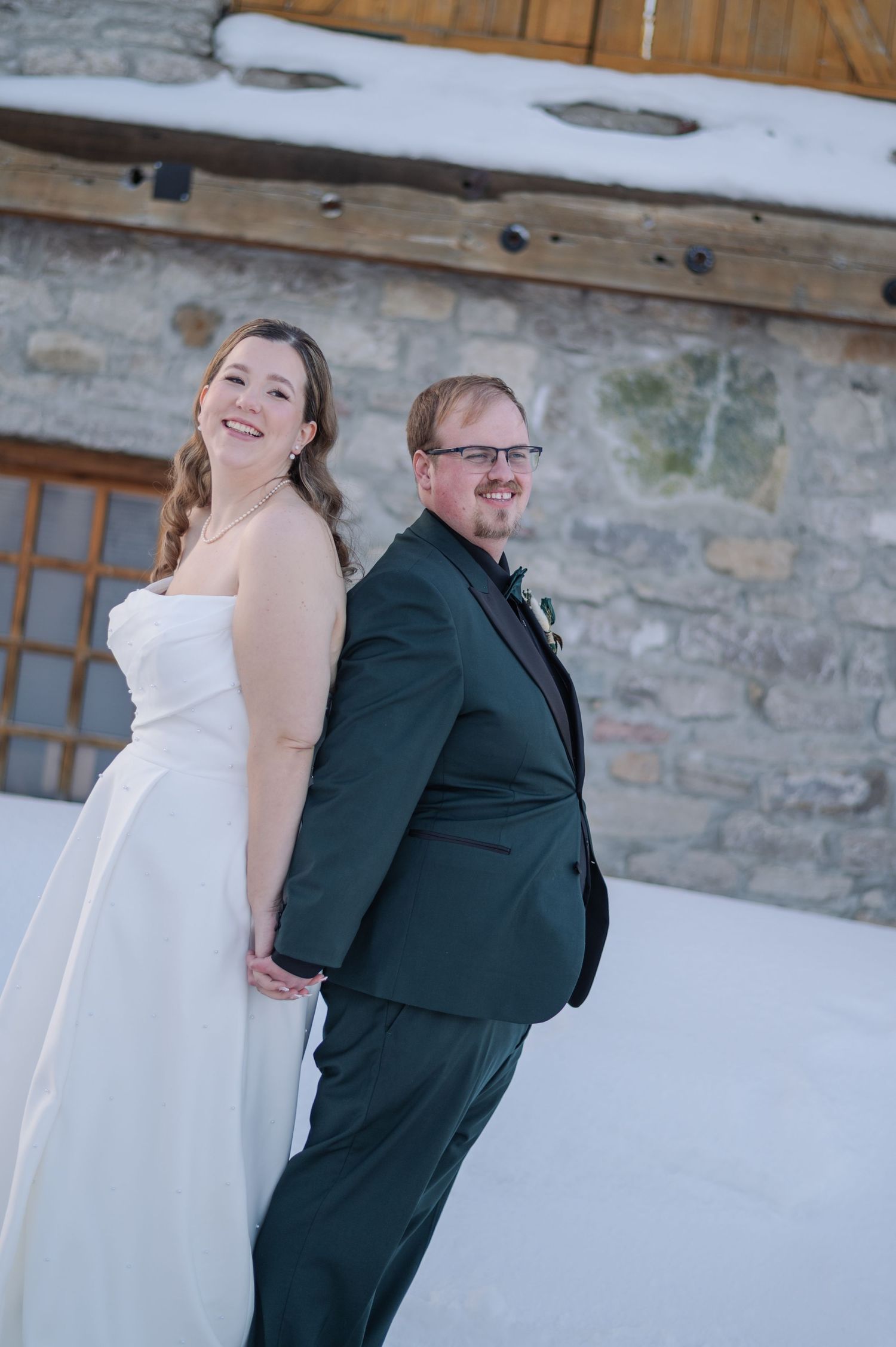 A bride in a white gown and groom in a dark green suit pose back-to-back in the snow outside a stone building.