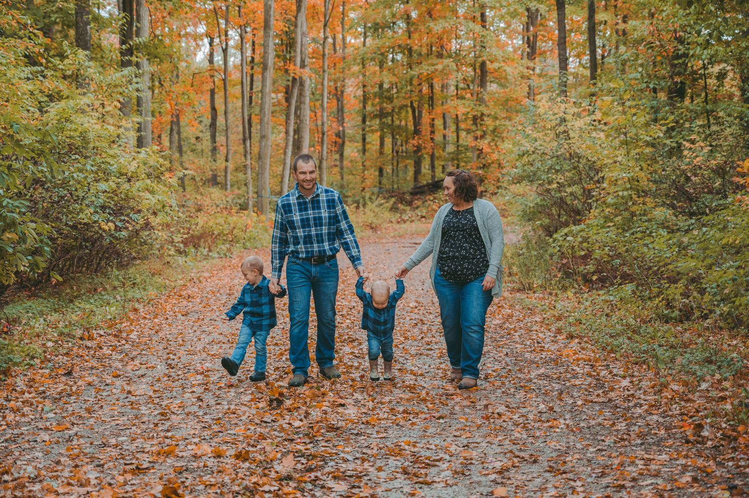 Family of four walking hand in hand down a scenic autumn forest trail surrounded by orange trees.