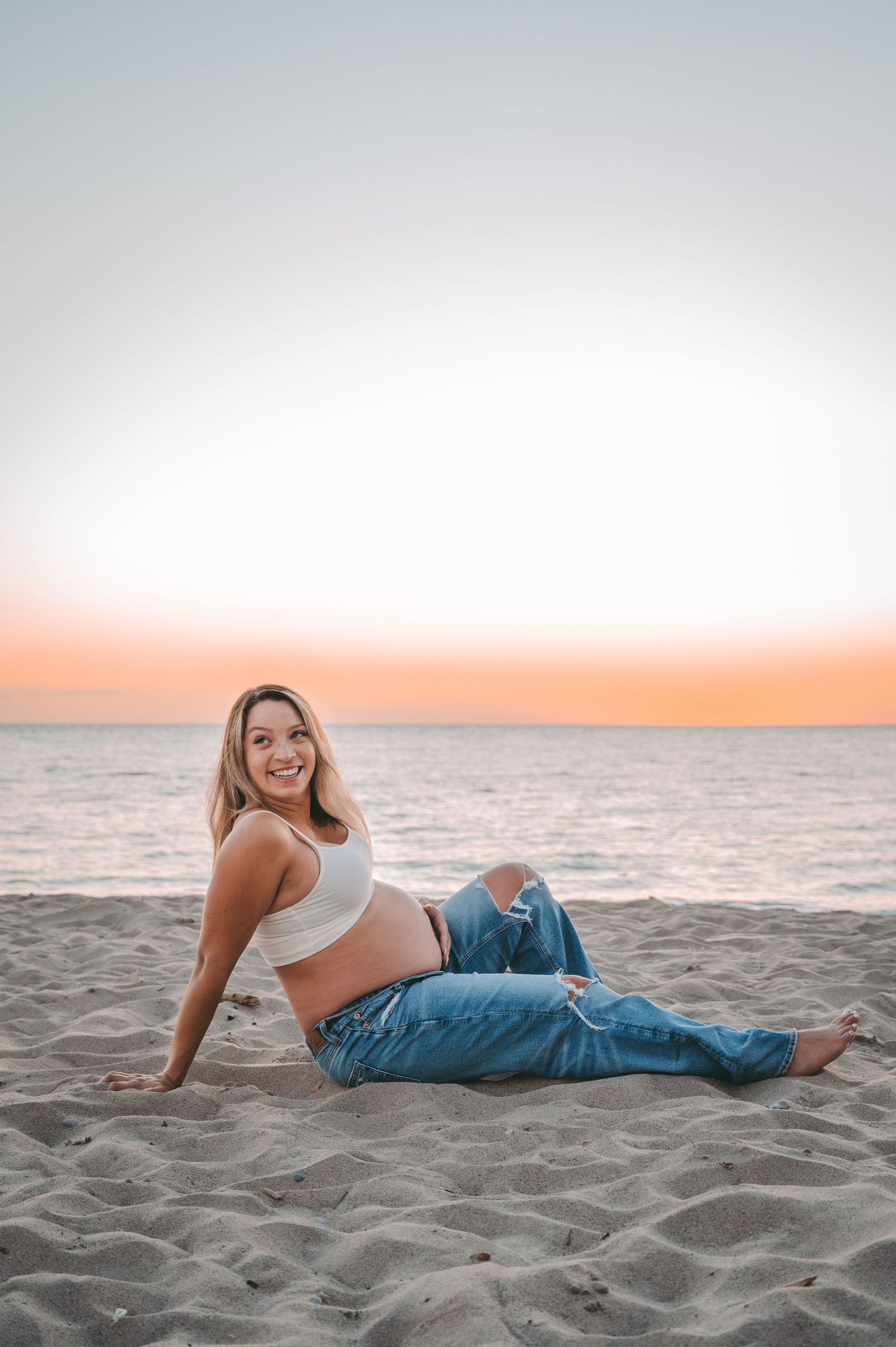 Someone wearing a white crop top and jeans lounges on a sandy beach at sunset with orange sky over the ocean.