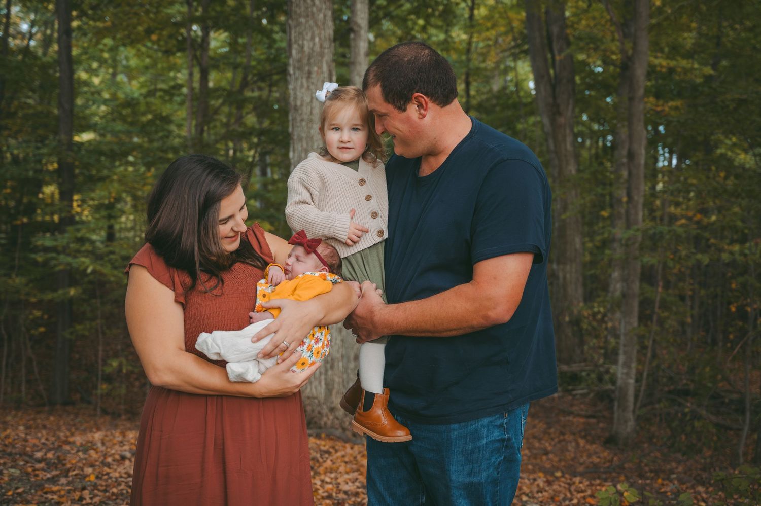 Family stands together in autumn forest setting holding baby and young child in heartwarming outdoor portrait.