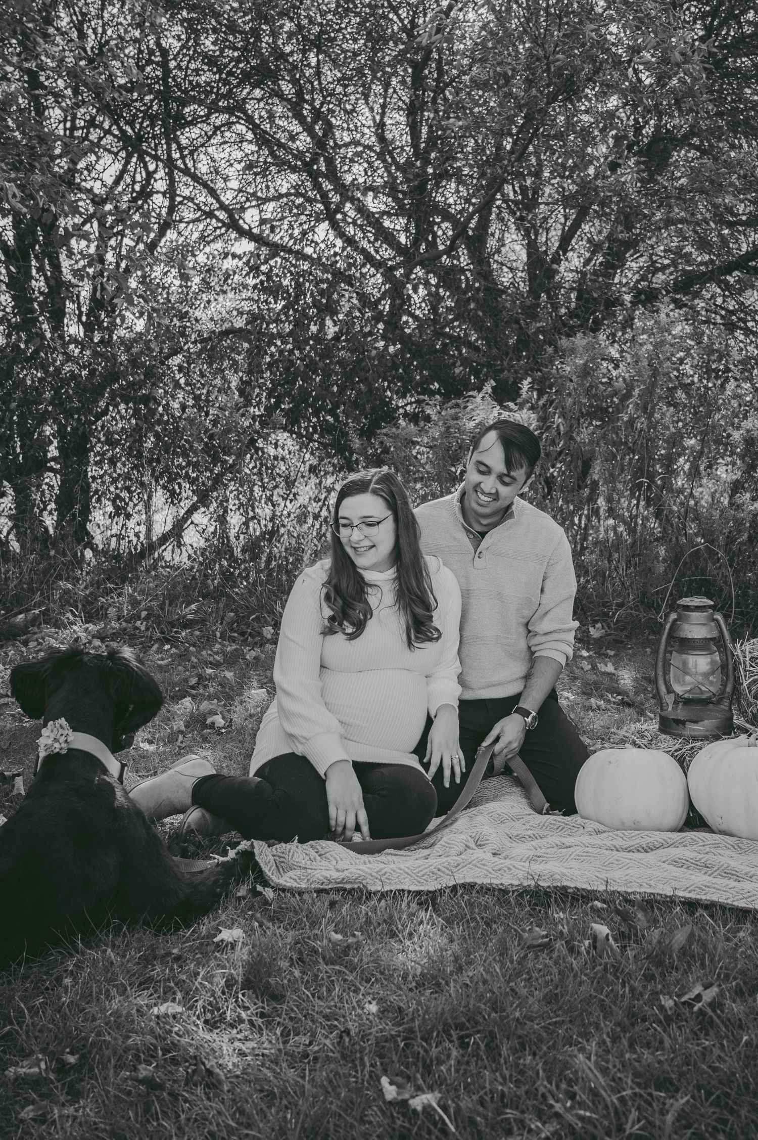 A couple enjoys an outdoor picnic with pumpkins and a lantern on a blanket under autumn trees.