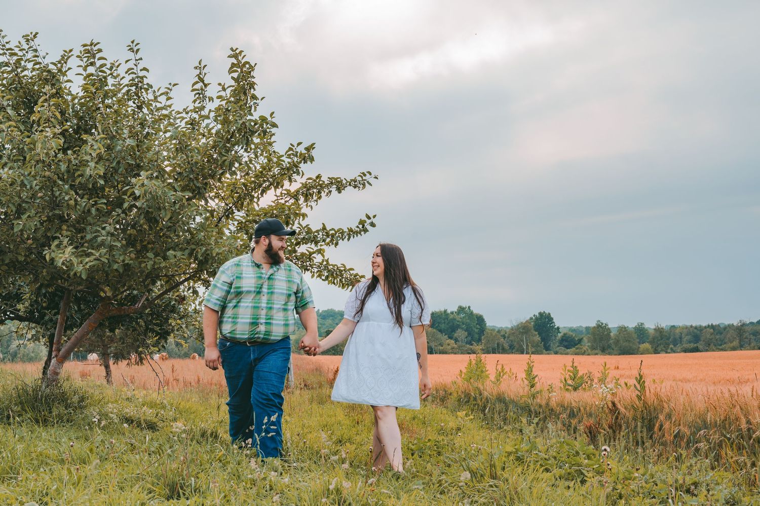 A couple holding hands while walking along the edge of an orchard near a wheat field.