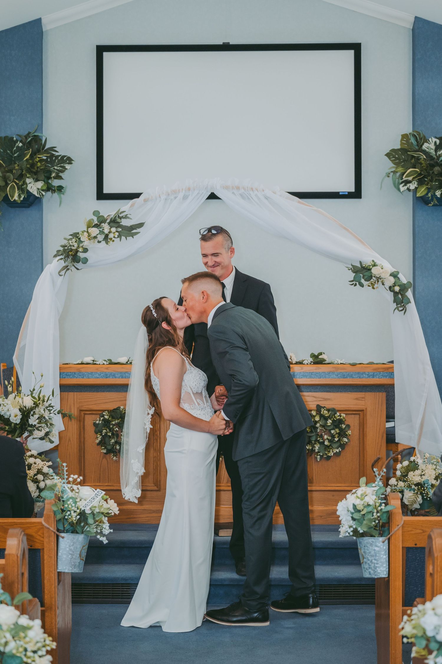 Wedding ceremony under a white arch decorated with greenery in a blue-walled church.