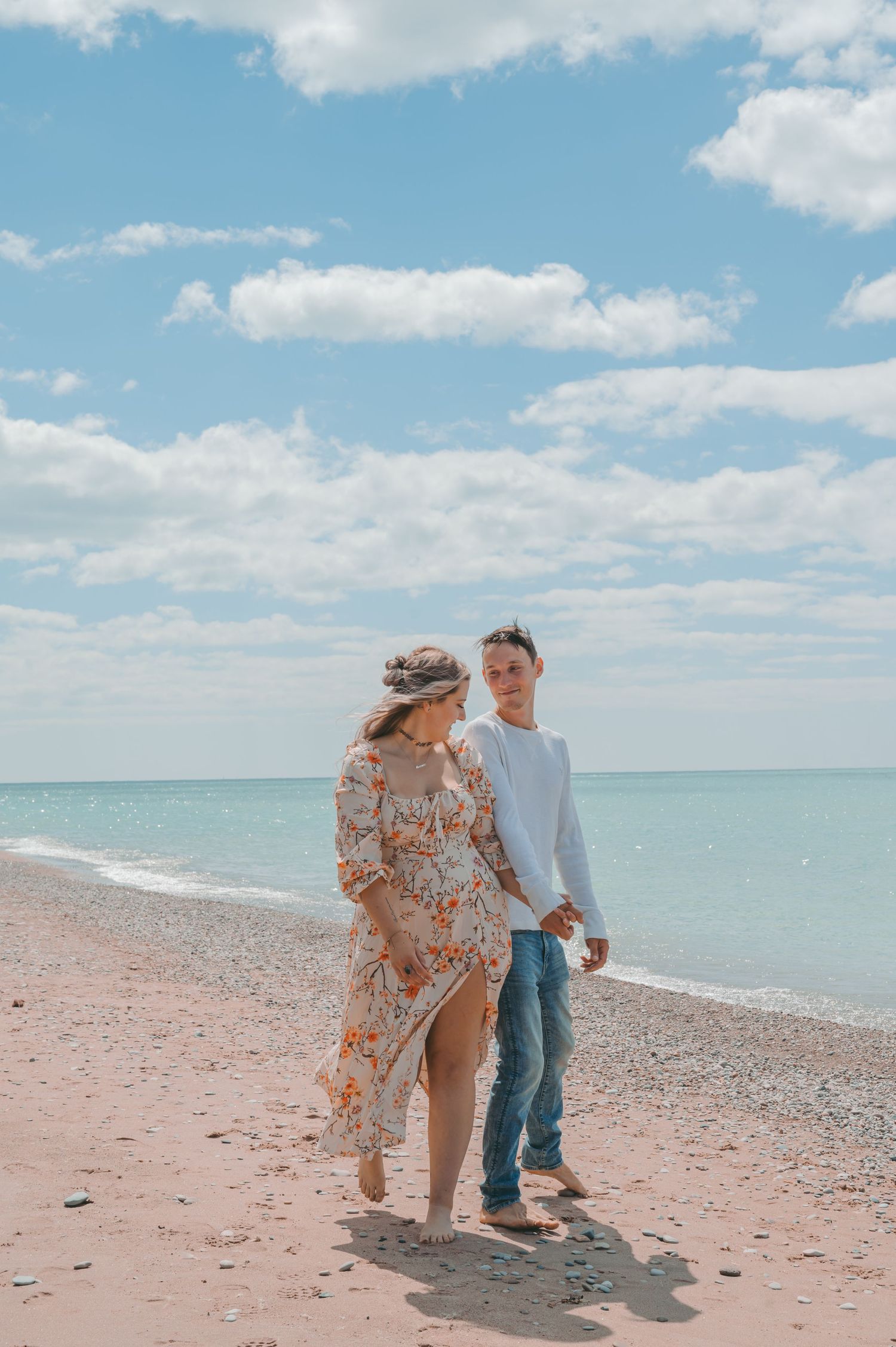 Couple walking on pebble beach with floral dress flowing in breeze against blue ocean backdrop.
