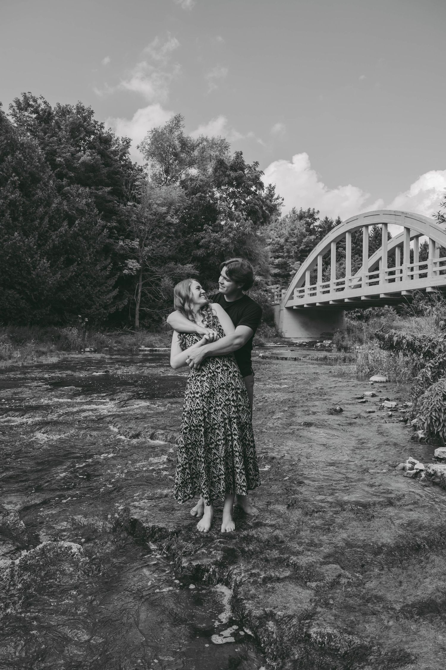 Black and white portrait of couple embracing by river with arched bridge backdrop.