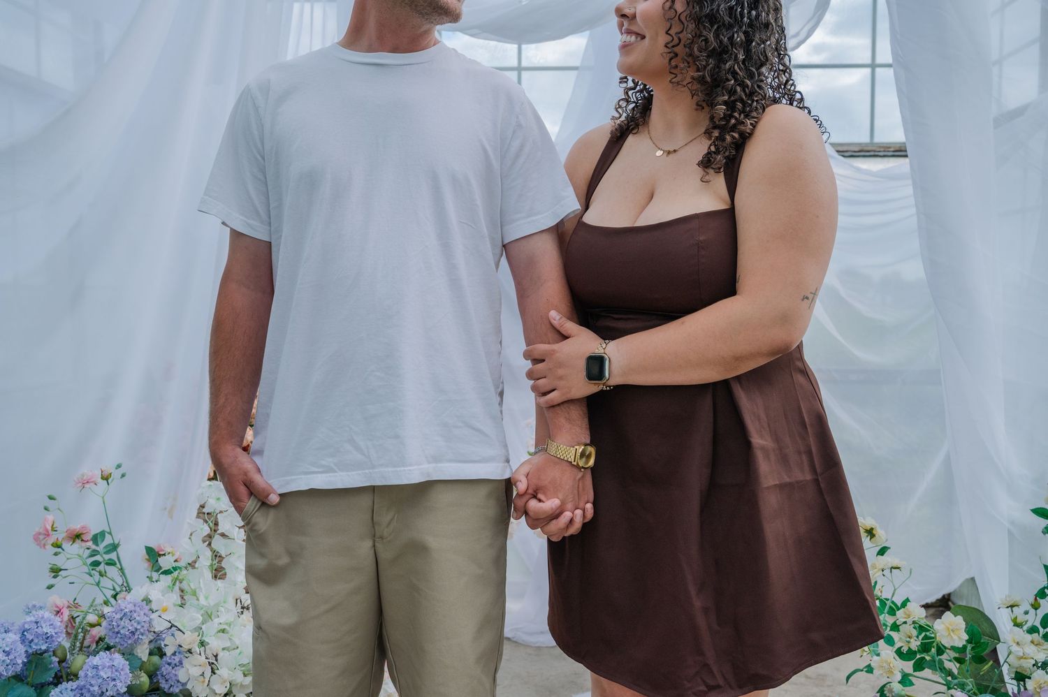 Couple holding hands in front of white drapes and floral decor, man in white tee and woman in brown mini dress.