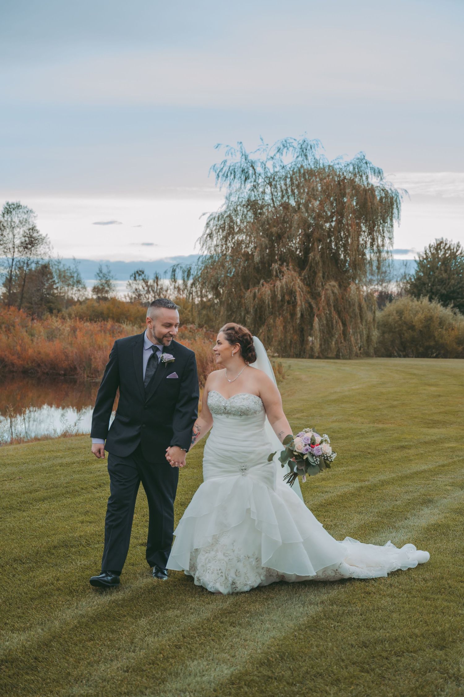 Wedding couple poses together on a green lawn with willow trees and pond in the background during autumn.
