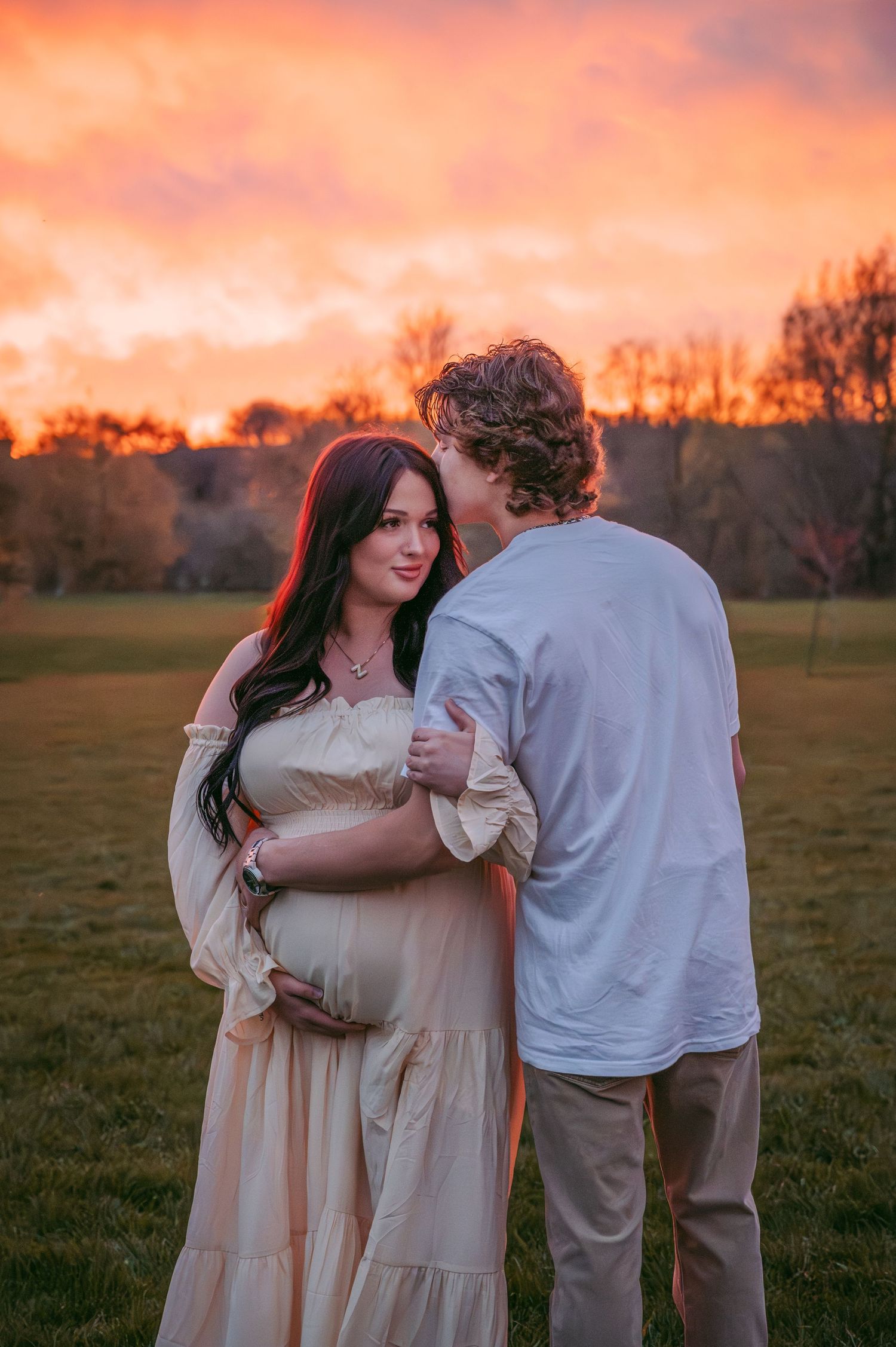 A couple embraces in a field at sunset with warm orange sky glowing behind them.