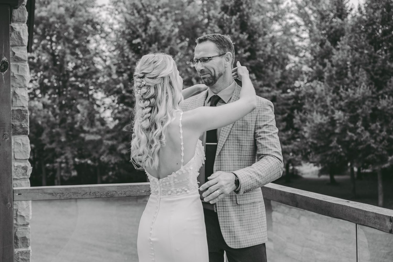 Sentimental black and white wedding photo of a father and daughter embracing on an outdoor deck surrounded by trees.