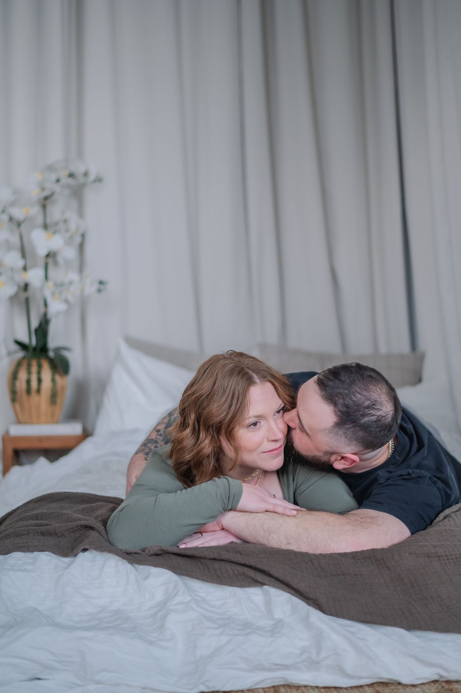 A couple shares a tender moment on a bed, the man kissing the smiling woman's cheek in a cozy bedroom.