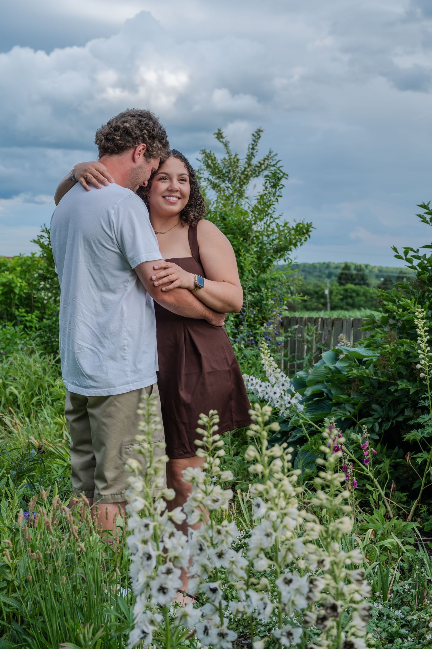 Couple embracing in a lush garden with white wildflowers, woman in brown dress smiling, cloudy sky backdrop.