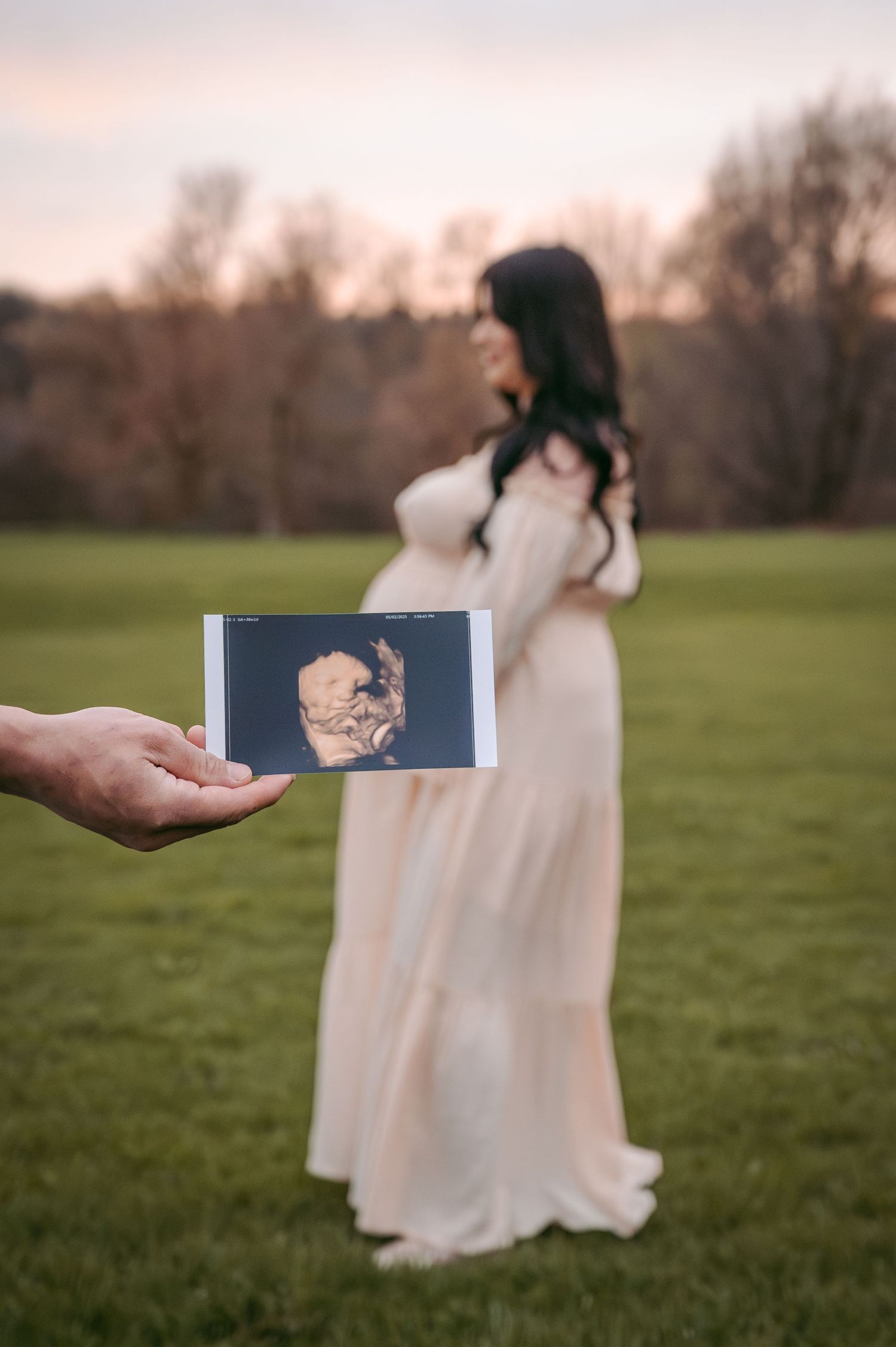 Person holding ultrasound photo while pregnant woman in white dress stands in background at sunset.