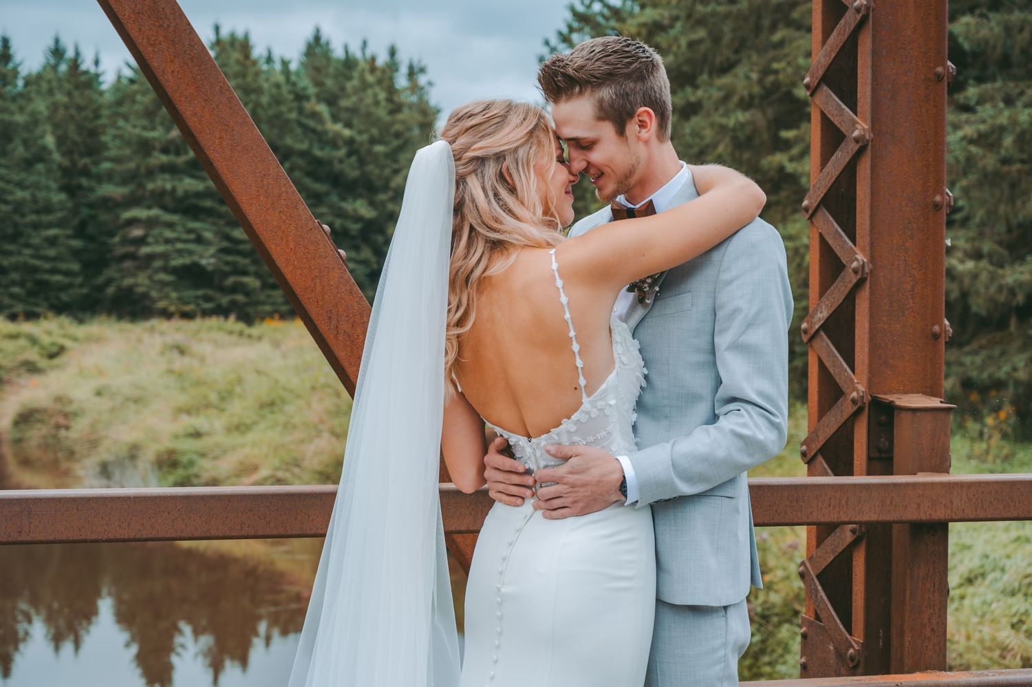 A romantic wedding couple embraces on a rustic wooden bridge over a pond with evergreen trees in the background.