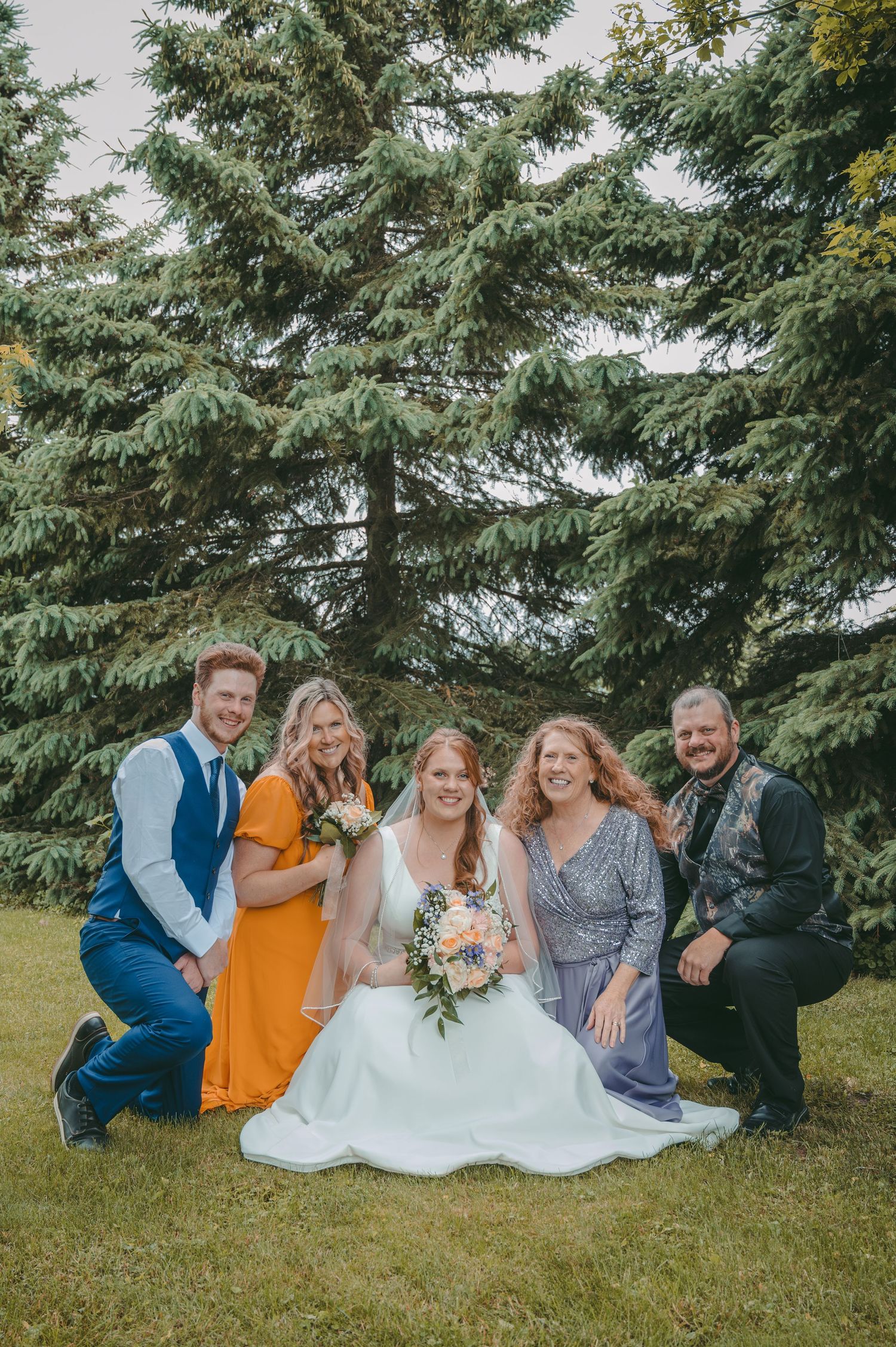 Family poses casually in front of evergreen trees with bride in white dress and bridesmaids in orange and blue.