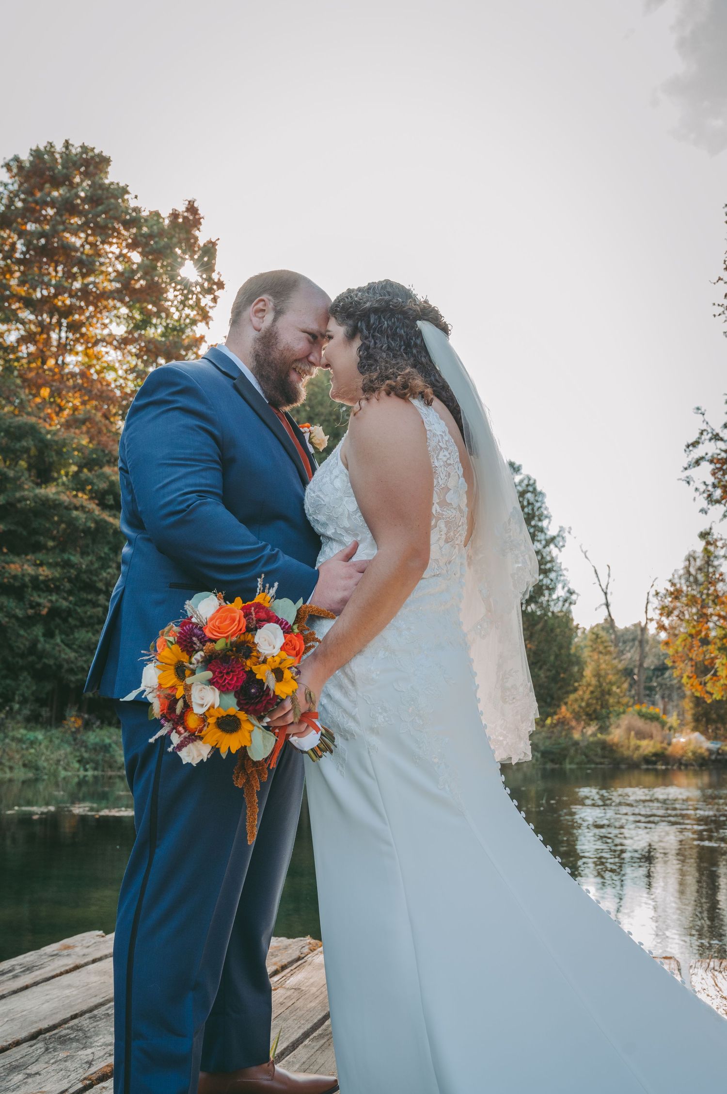 Bride and groom pose with orange bouquet on wooden dock by lake during fall wedding.